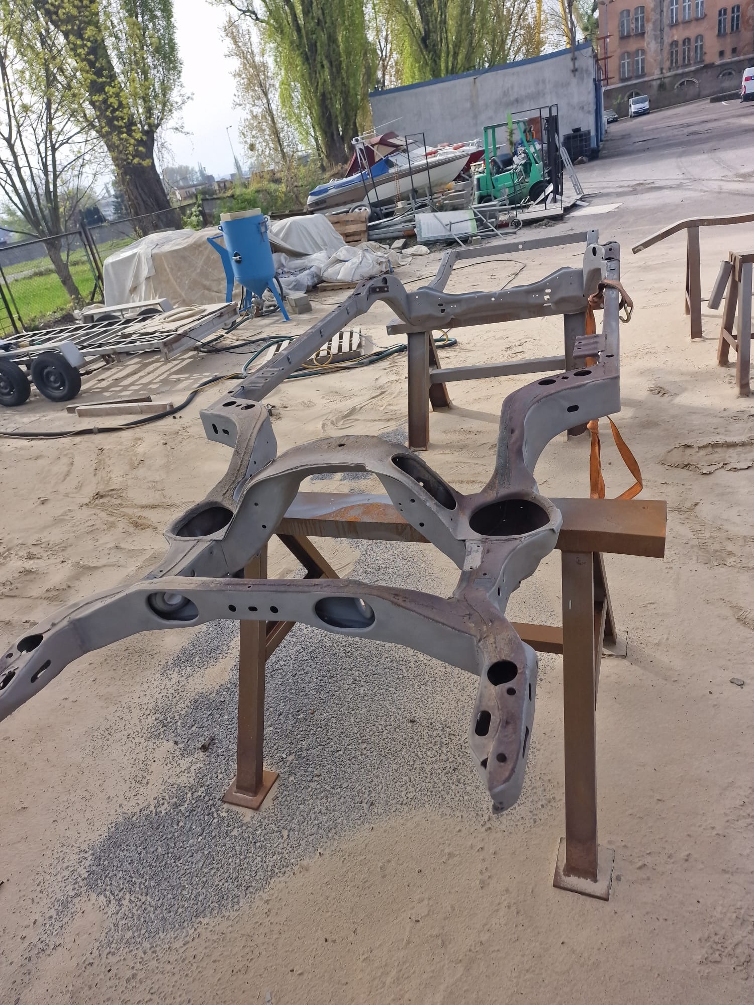 Szara, metalowa rama samochodowa, częściowo oczyszczona, leży na dwóch stalowych kozłach na zewnątrz, w tle widoczne inne elementy i budynki.