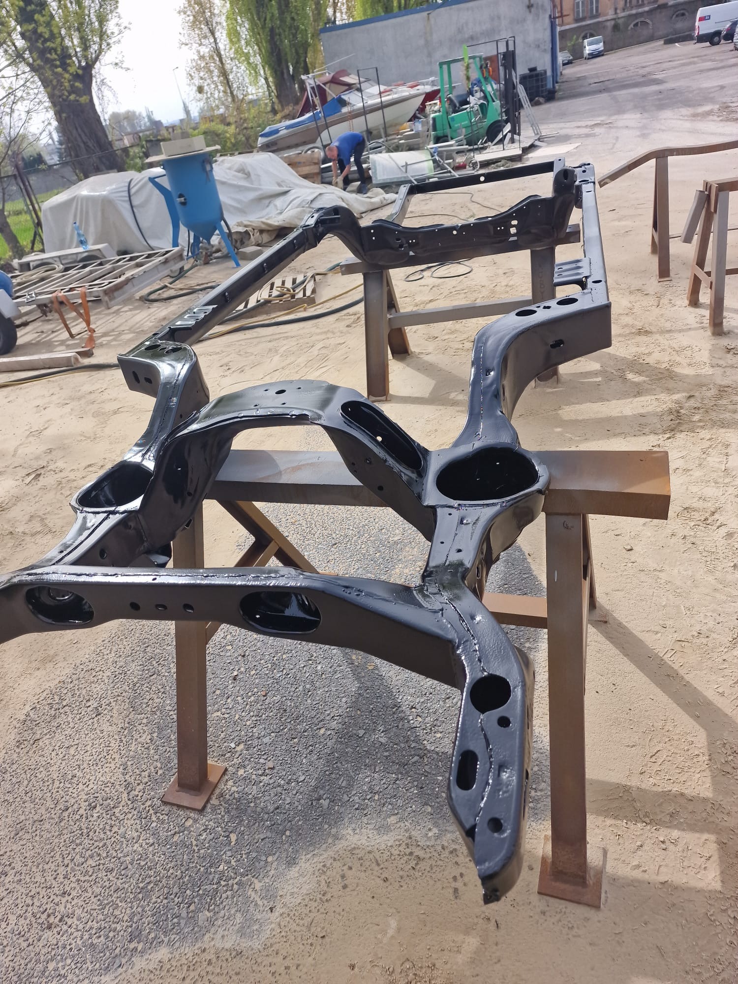 Błyszcząca, czarna rama samochodu po renowacji, umieszczona na metalowych kozłach w warsztacie, z widocznym sprzętem do piaskowania i pracującym mężczyzną w tle.