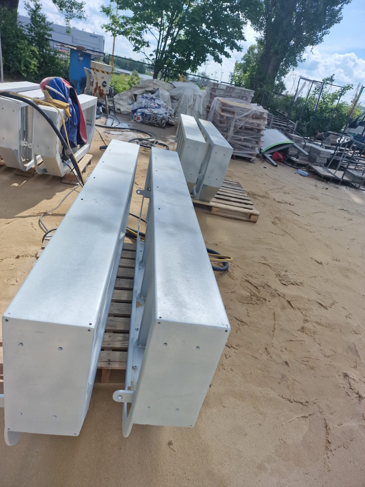 Dwa elementy konstrukcyjne pomalowane na szaro, leżące na paletach na piaszczystym podłożu, prawdopodobnie po piaskowaniu i malowaniu proszkowym.