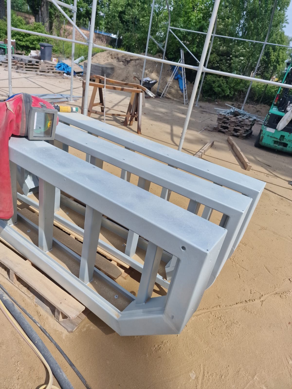 Pomalowane na szaro metalowe ramy konstrukcyjne, leżące na palecie na piaszczystym podłożu, z widocznym sprzętem budowlanym w tle i maską spawalniczą.