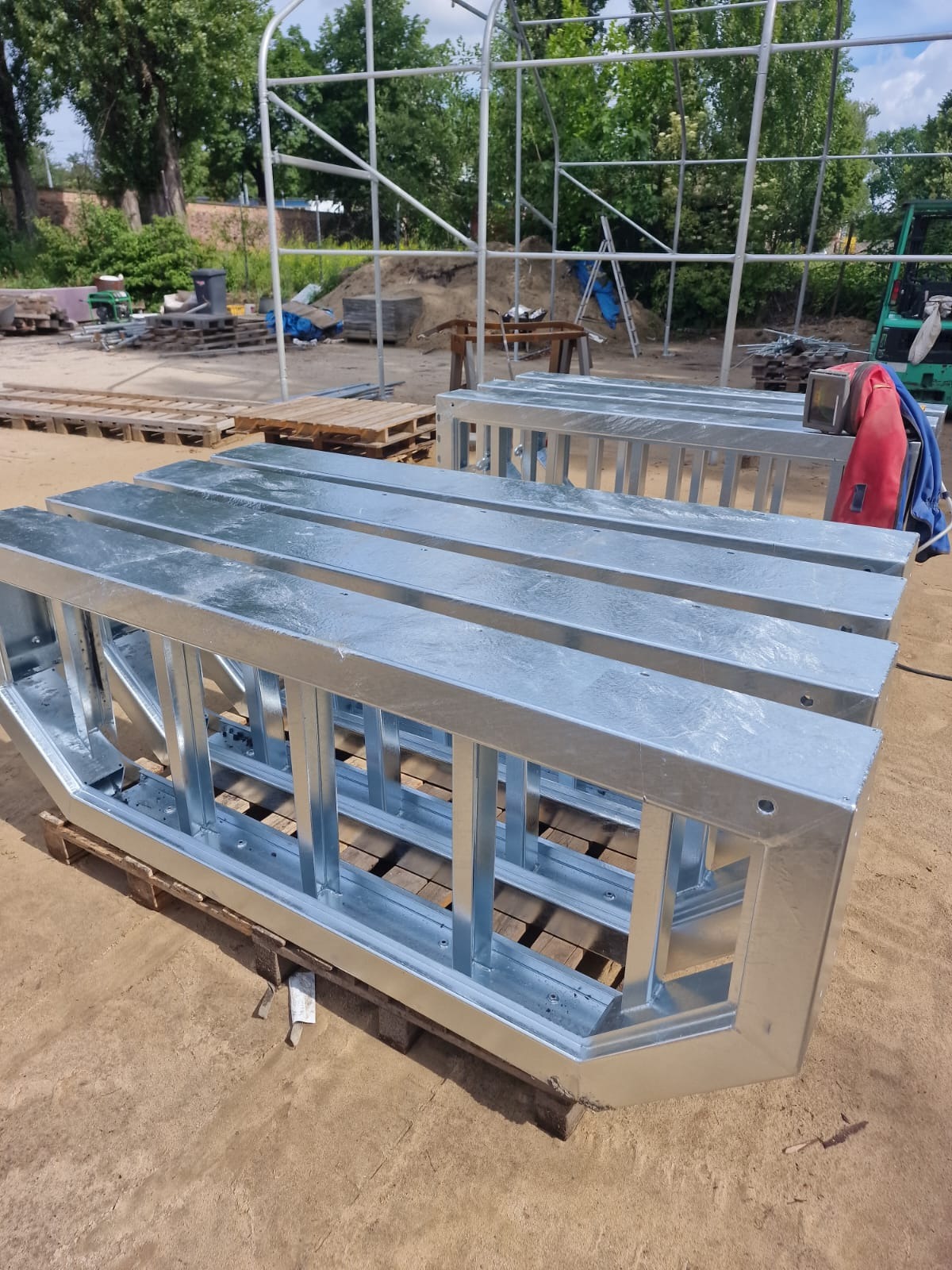 Metalowe, ocynkowane elementy konstrukcyjne, prawdopodobnie ramy, ułożone na paletach na zewnątrz, w tle widoczna konstrukcja szkieletowa i sprzęt budowlany.