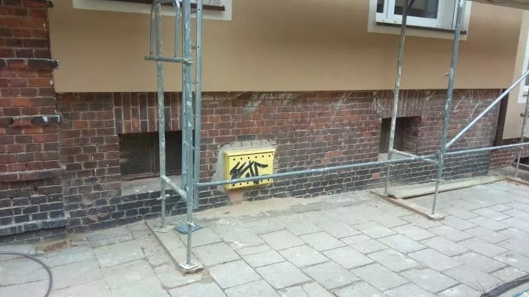 Elewacja budynku po częściowym ociepleniu z widocznym fragmentem rusztowania, ceglaną podmurówką i żółtą skrzynką gazową z czarnymi bazgrołami.