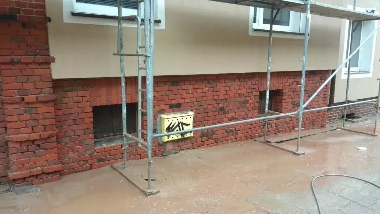 Ceglana elewacja budynku w trakcie renowacji, z widocznymi rusztowaniami, żółtą skrzynką z graffiti i mokrym chodnikiem.