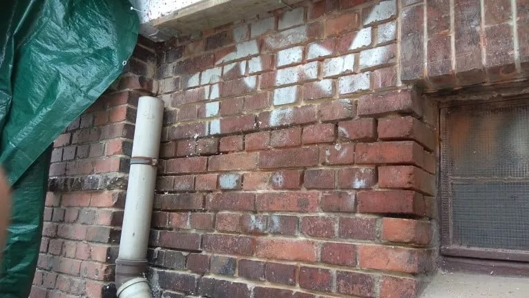 Ceglana ściana budynku z widoczną rurą kanalizacyjną, częściowo zasłonięta zieloną plandeką, z białymi śladami po farbie na cegłach i oknem z kratą w tle.