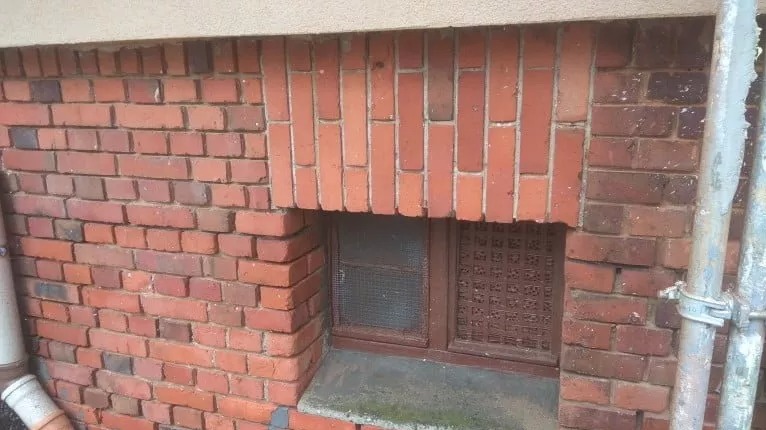 Zamknięte okienko piwniczne z kratką, wmurowane w ceglany mur budynku, z widoczną rurą spustową po lewej stronie i fragmentem rusztowania po prawej.
