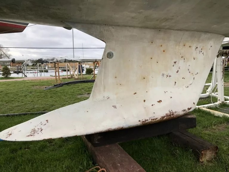 Dolna część kadłuba łodzi z widocznymi śladami korozji i ubytkami farby, wsparta na drewnianych belkach na trawie, z fragmentem mariny w tle.