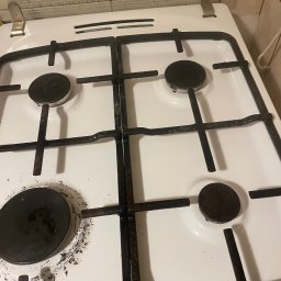 Clover - Zabrudzona, biała kuchenka gazowa z czarnymi palnikami i rusztem, widoczne ślady przypaleń i zaschniętego brudu, górna klapa z instrukcjami.