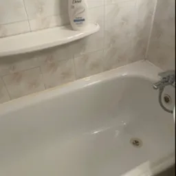 Biała, pusta wanna z chromowaną baterią i odpływem, na półce butelka mydła Dove, widok z góry, płytki w tle.