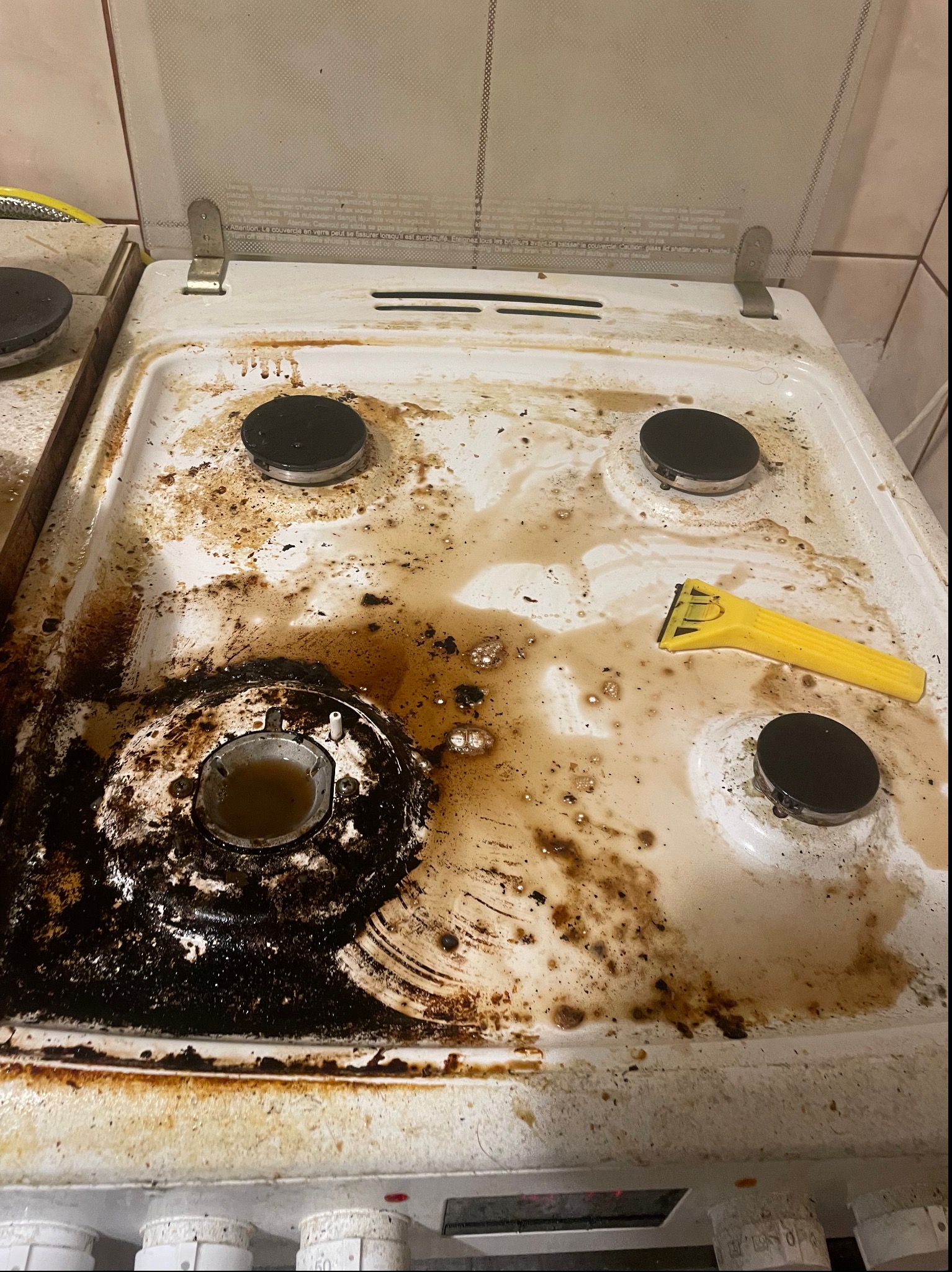 Zabrudzona, biała płyta kuchenna z czarnymi palnikami, resztkami jedzenia i żółtą szpachelką. Jeden z palników jest zalany brudną wodą.