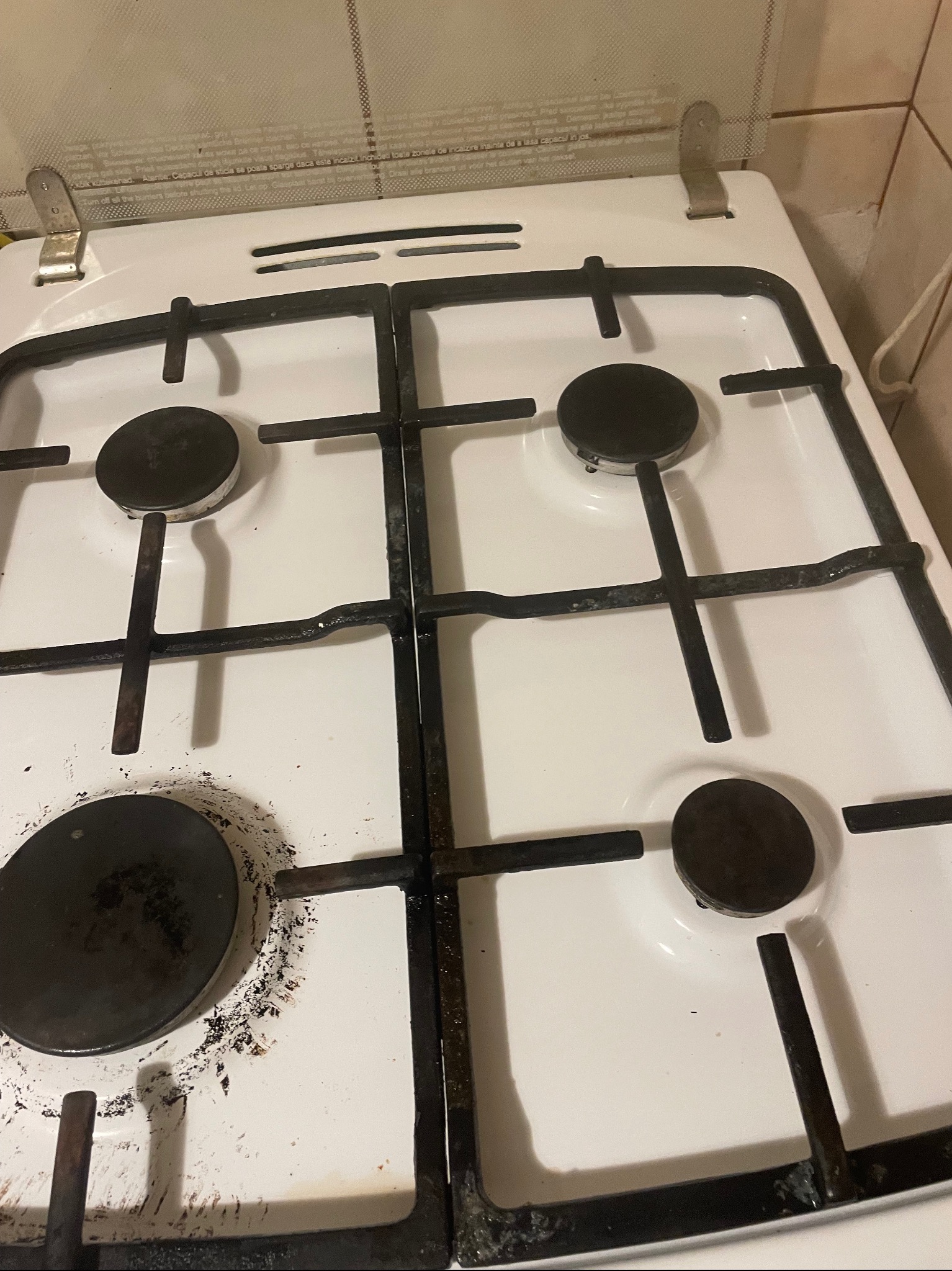 Zabrudzona, biała kuchenka gazowa z czarnymi palnikami i rusztem, widoczne ślady przypaleń i zaschniętego brudu, górna klapa z instrukcjami.