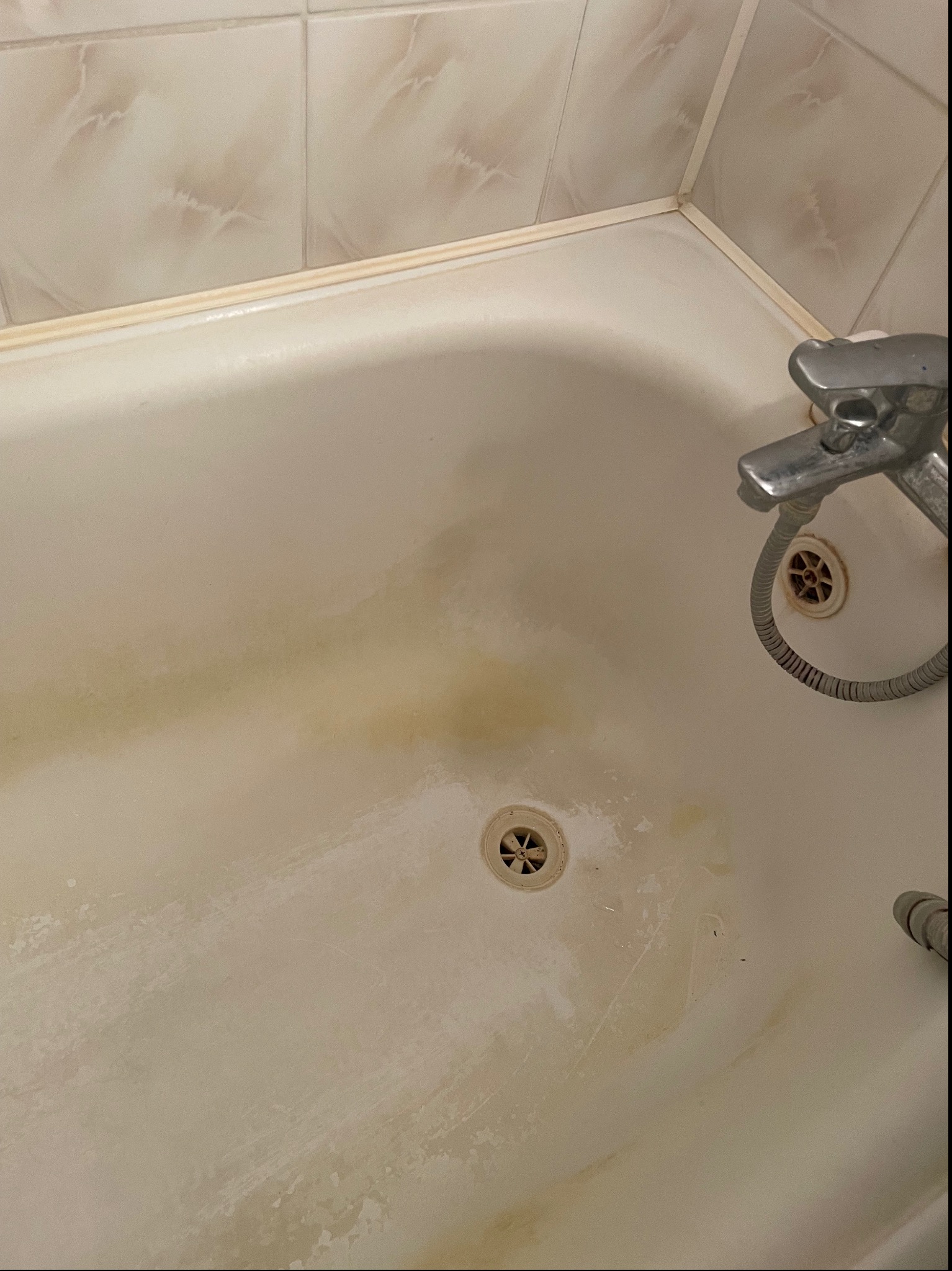 Zabrudzona, biała wanna z żółtymi zaciekami, widoczne dwa odpływy, bateria i słuchawka prysznicowa.