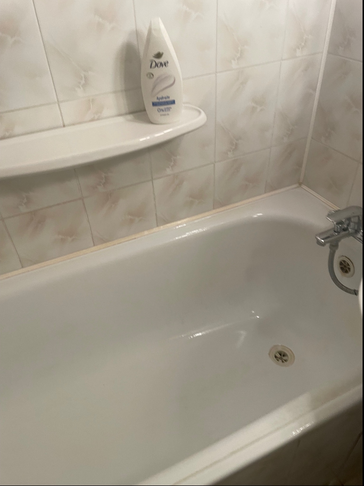 Biała, pusta wanna z chromowaną baterią i odpływem, na półce butelka mydła Dove, widok z góry, płytki w tle.