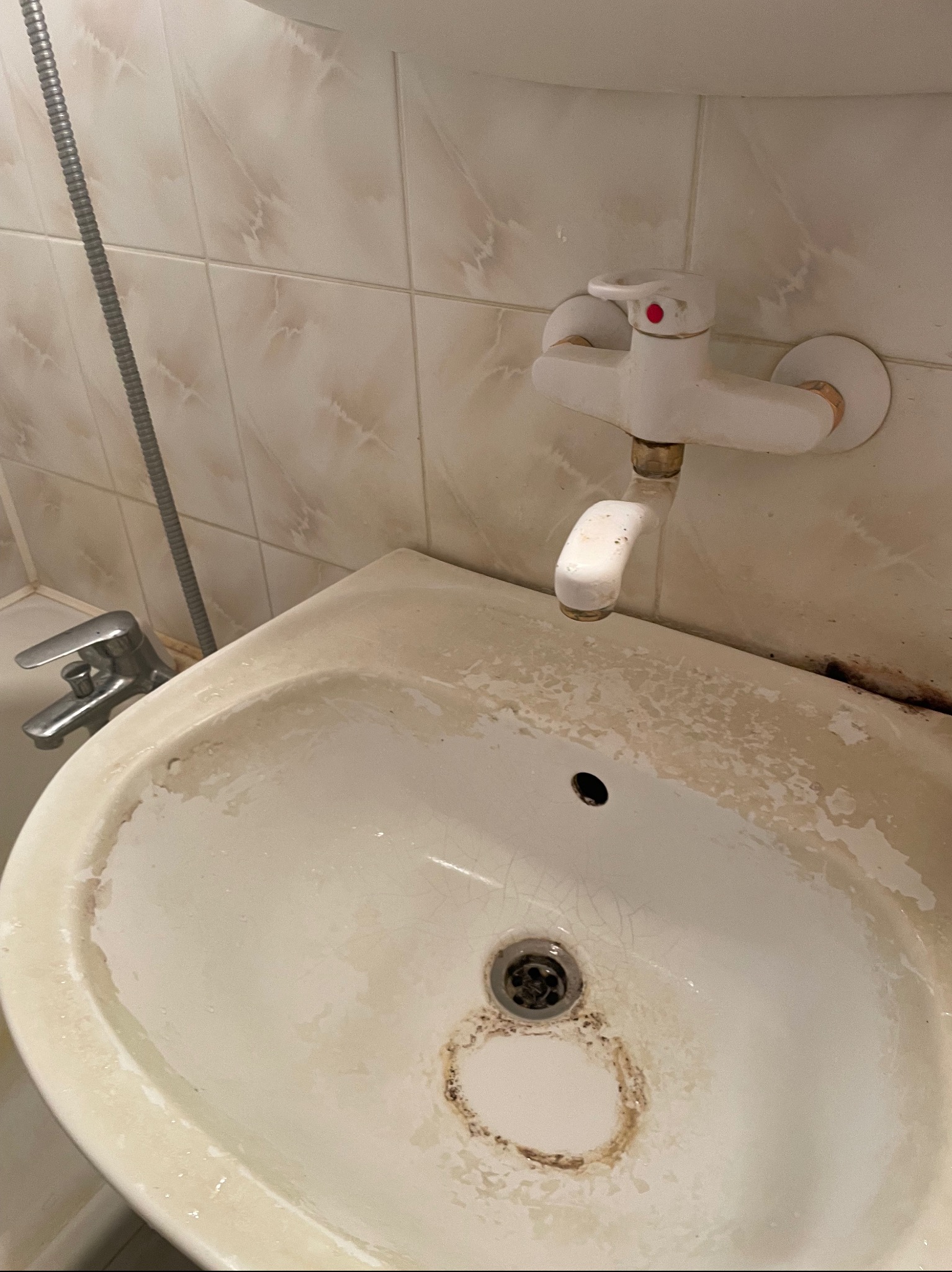 Zabrudzona umywalka z białym kranem i śladami pleśni w łazience z beżowymi płytkami.