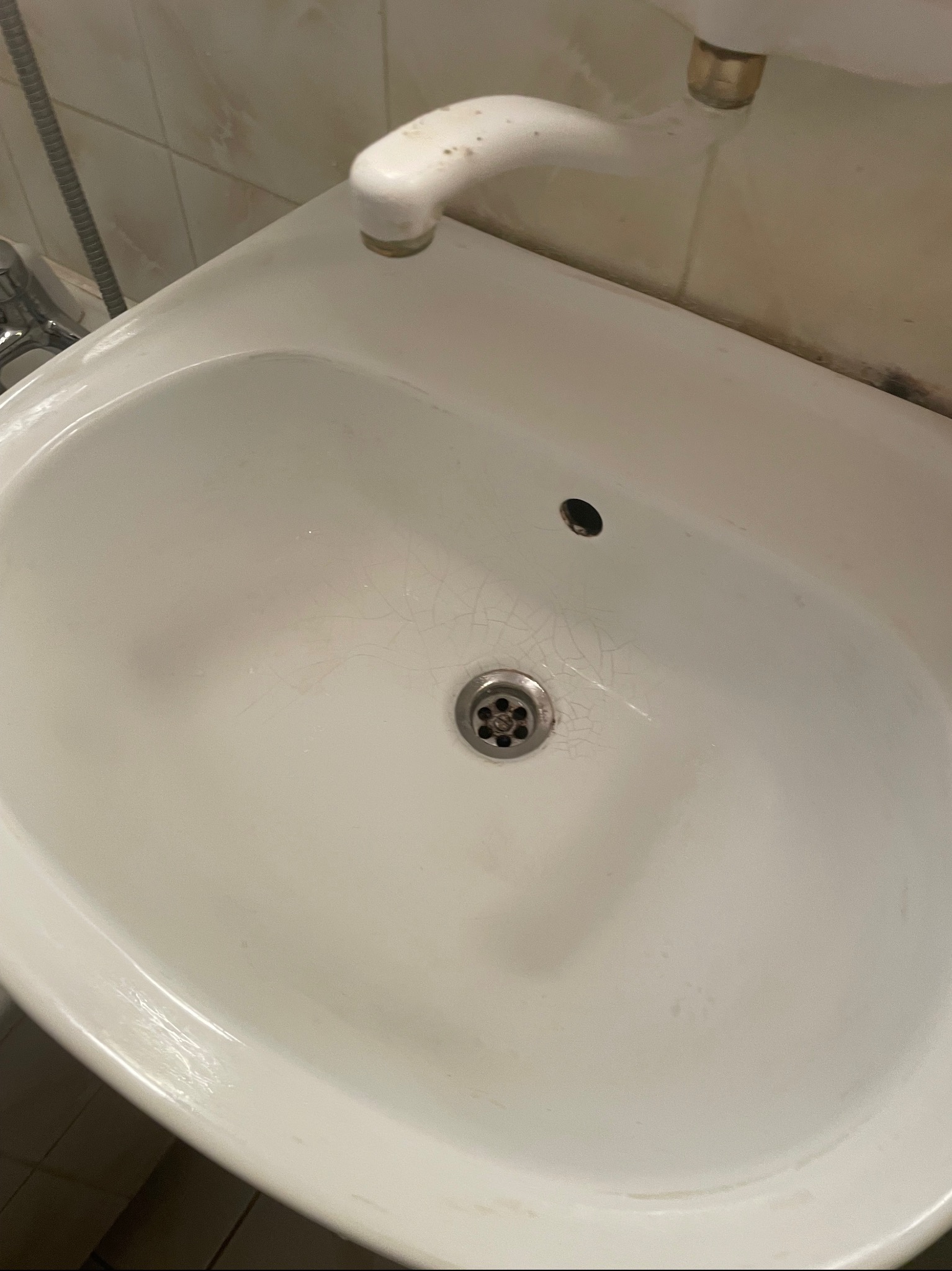 Biała, ceramiczna umywalka z widocznymi pęknięciami i zabrudzeniami, z kranem i odpływem, sfotografowana z góry.