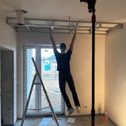 DOM-BUD Dominik Budyś - Montaż stelaża sufitu podwieszanego z płyty gipsowej. Widoczny pracownik, drabina i narzędzia. W tle okno z widokiem na zewnątrz. Wnętrze w stanie surowym.