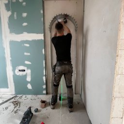 DOM-BUD Dominik Budyś - Pracownik montuje metalową konstrukcję łukową do ściany z płyt karton-gips, używając wkrętarki. Widoczne narzędzia i laserowy poziom. W tle surowe ściany w trakcie prac.