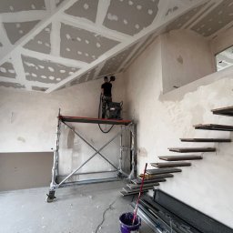 DOM-BUD Dominik Budyś - Pracownik szlifuje gładź na poddaszu z rusztowania. Widoczne płyty karton-gips na suficie, schody wiszące i narzędzia malarskie. Wnętrze w trakcie wykończenia.