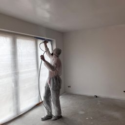DOM-BUD Dominik Budyś - Pracownik w kombinezonie ochronnym maluje ściany i sufit w pomieszczeniu za pomocą pistoletu natryskowego. Okno zabezpieczone folią malarską.