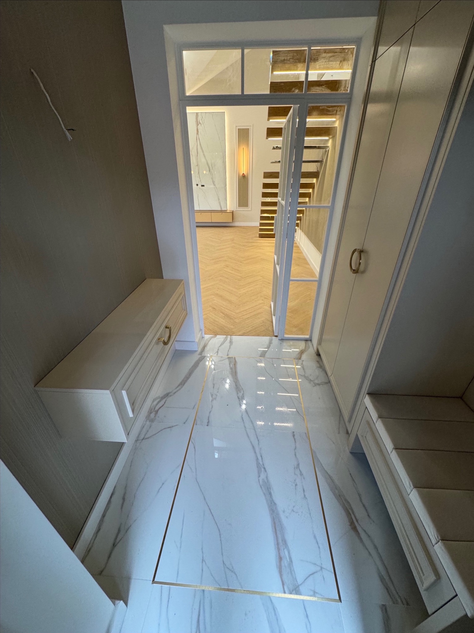 Biała marmurowa podłoga z złotymi akcentami, widok na schody z oświetleniem LED. Eleganckie wykończenie wnętrza z szafkami w zabudowie i otwartym przejściem.