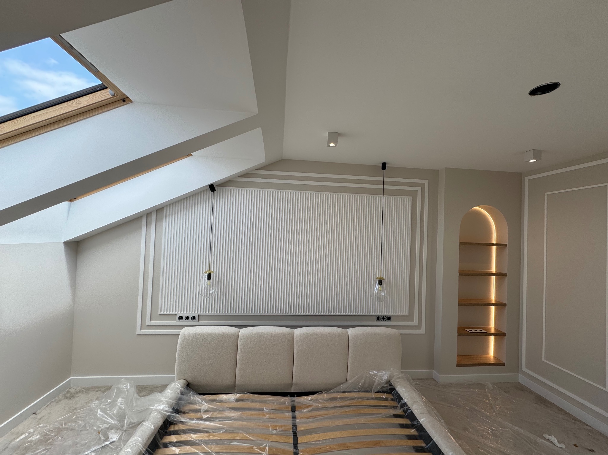 Pokój na poddaszu z łóżkiem w folii, dekoracyjną ścianą z pionowymi żłobieniami, oświetloną wnęką i oknem dachowym. Minimalistyczny design w jasnych barwach.