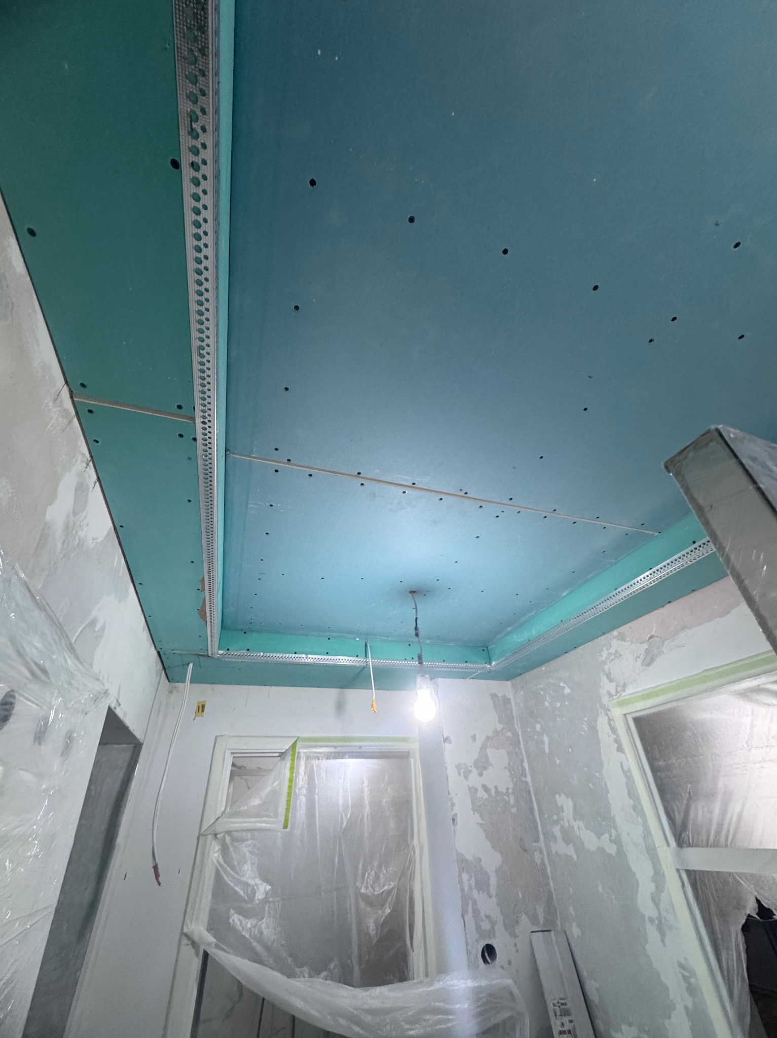 Niebieskie płyty gipsowe zamontowane na suficie z metalowymi profilami, widok z dołu. Okna zabezpieczone folią malarską. Etap prac wykończeniowych.