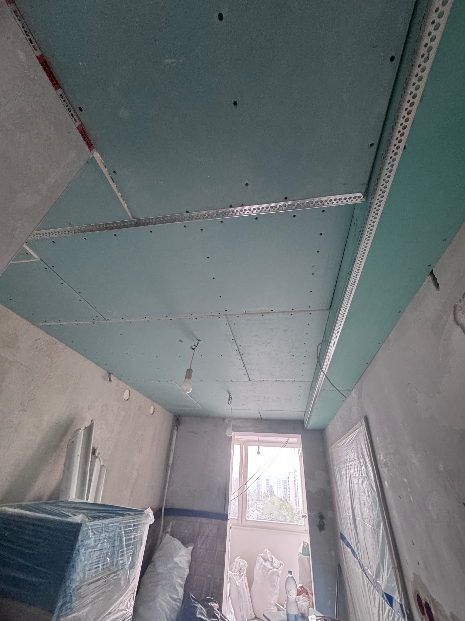Montaż zielonych płyt karton-gips na suficie z profilami aluminiowymi w surowym wnętrzu. Widoczne worki z materiałami budowlanymi i okno w tle.