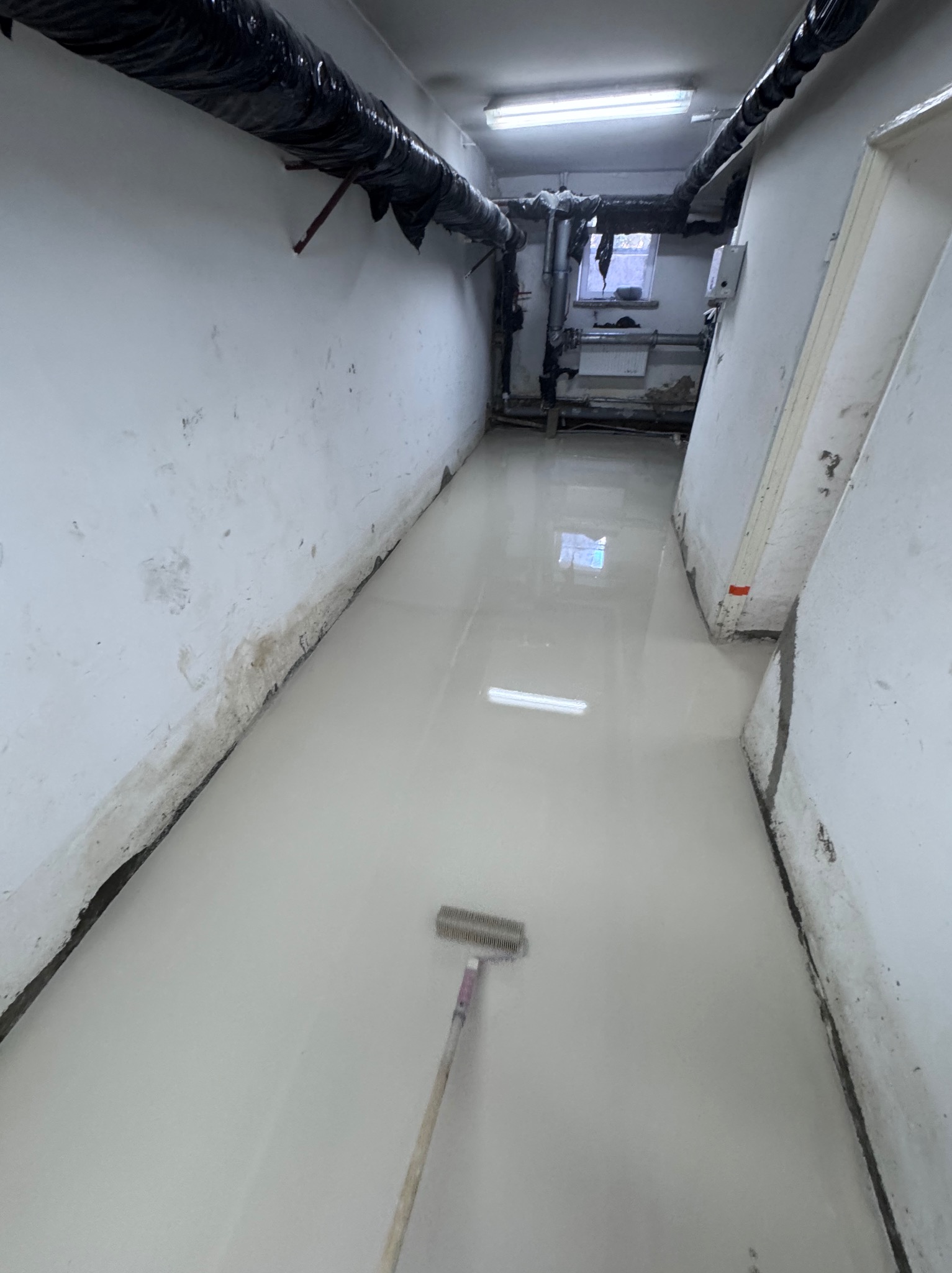 Wylewka samopoziomująca w piwnicy, widoczny pędzel do rozprowadzania masy. Rury pod sufitem. Okno w tle. Ściany w stanie surowym. Jasne, sztuczne oświetlenie.