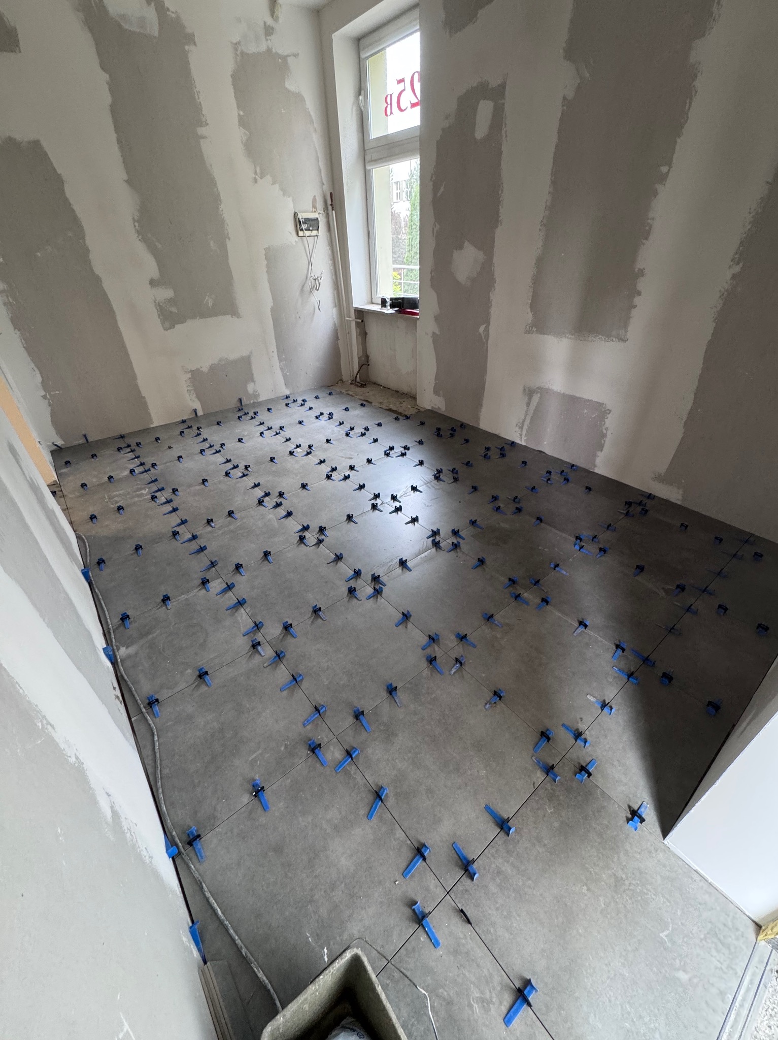 Szare płytki podłogowe z niebieskimi krzyżykami dystansowymi w trakcie układania. Ściany w surowym stanie, przygotowane do dalszych prac wykończeniowych. Widok z góry.