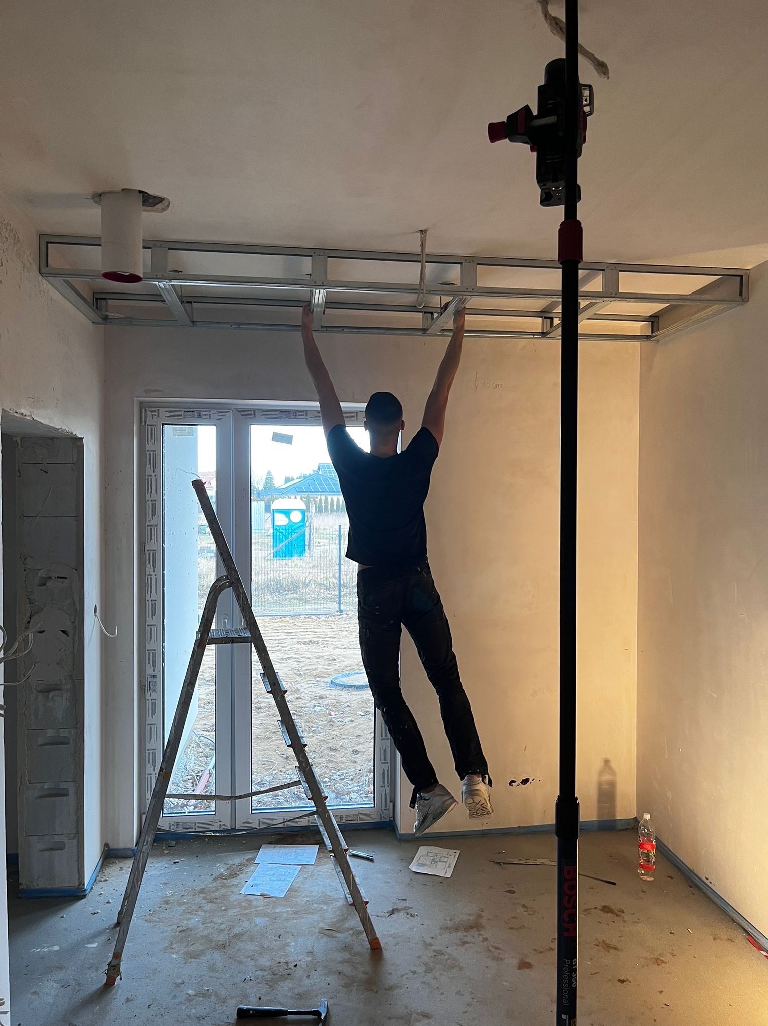 Montaż stelaża sufitu podwieszanego z płyty gipsowej. Widoczny pracownik, drabina i narzędzia. W tle okno z widokiem na zewnątrz. Wnętrze w stanie surowym.