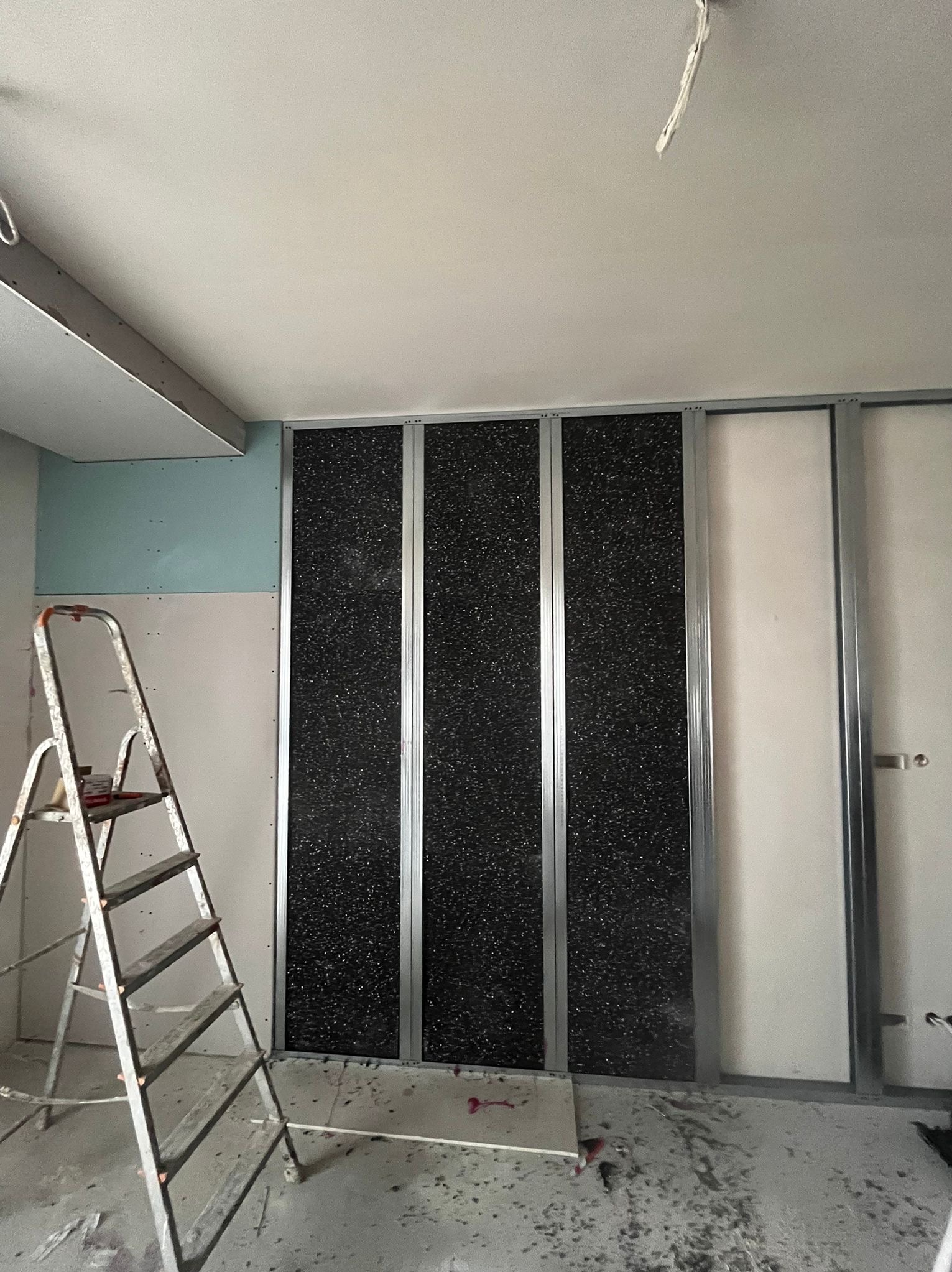 Montaż paneli ściennych z czarną, teksturowaną powierzchnią, widoczne metalowe profile i drabina. Prace wykończeniowe w surowym wnętrzu.