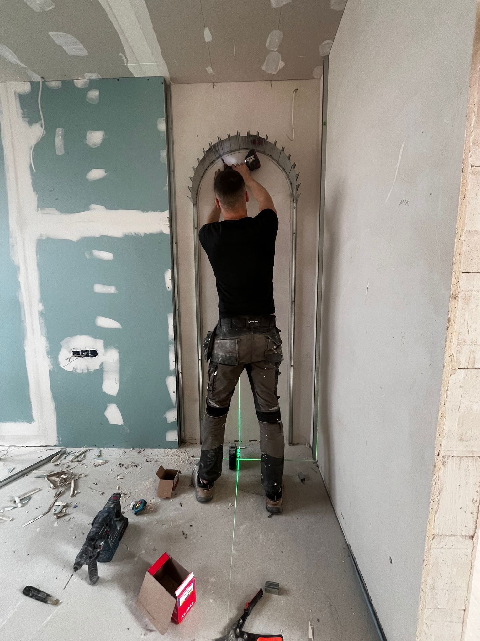 Pracownik montuje metalową konstrukcję łukową do ściany z płyt karton-gips, używając wkrętarki. Widoczne narzędzia i laserowy poziom. W tle surowe ściany w trakcie prac.