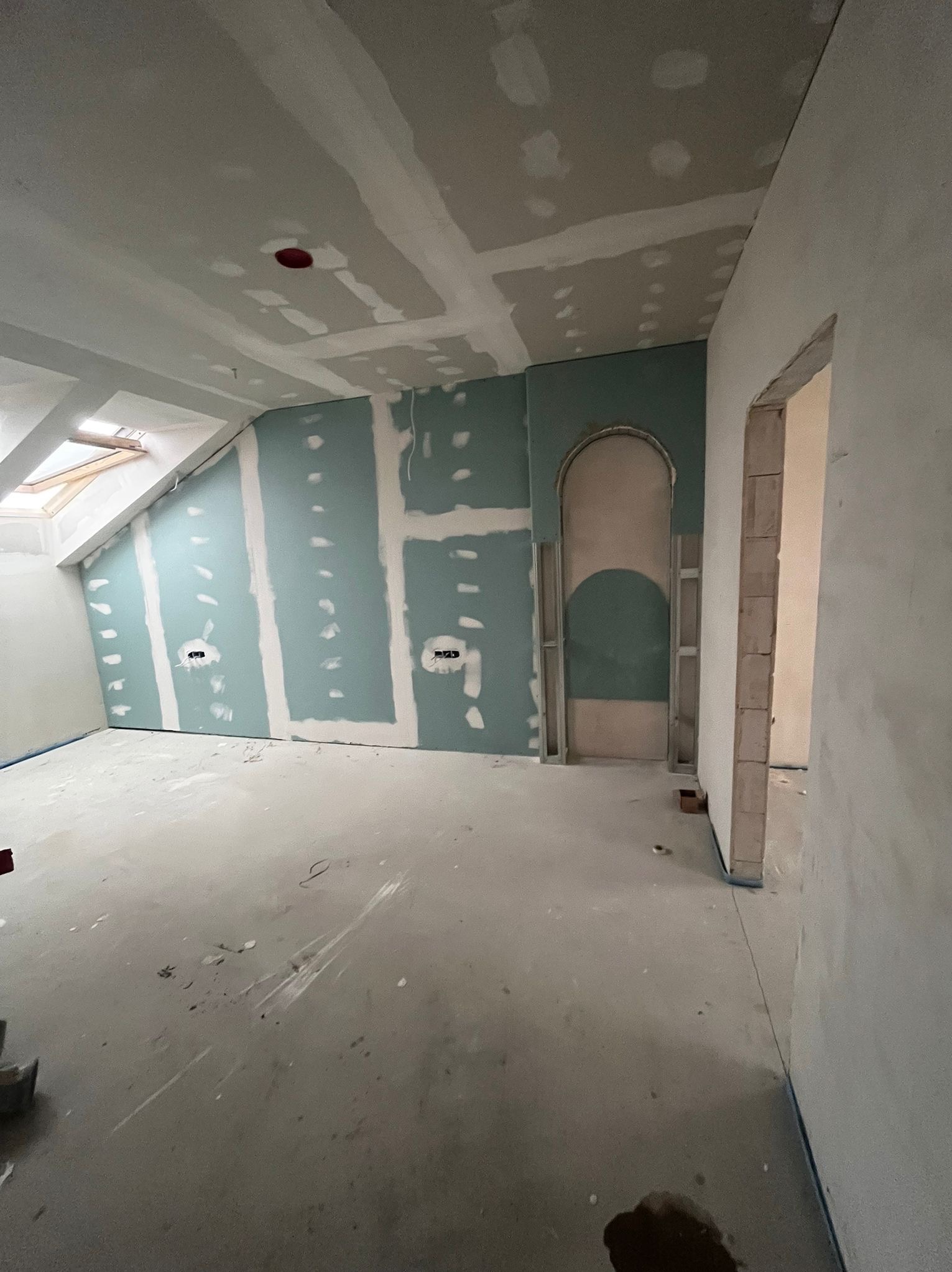 Poddasze w trakcie prac: ściany z płyt gipsowo-kartonowych, widoczne łączenia i otwory na gniazdka. Nietypowe łukowe przejście w ścianie. Okno dachowe w skosie.