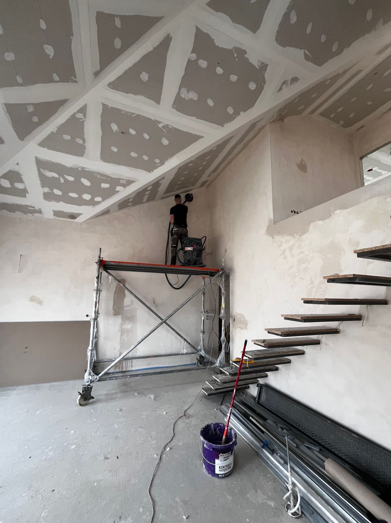 Pracownik szlifuje gładź na poddaszu z rusztowania. Widoczne płyty karton-gips na suficie, schody wiszące i narzędzia malarskie. Wnętrze w trakcie wykończenia.