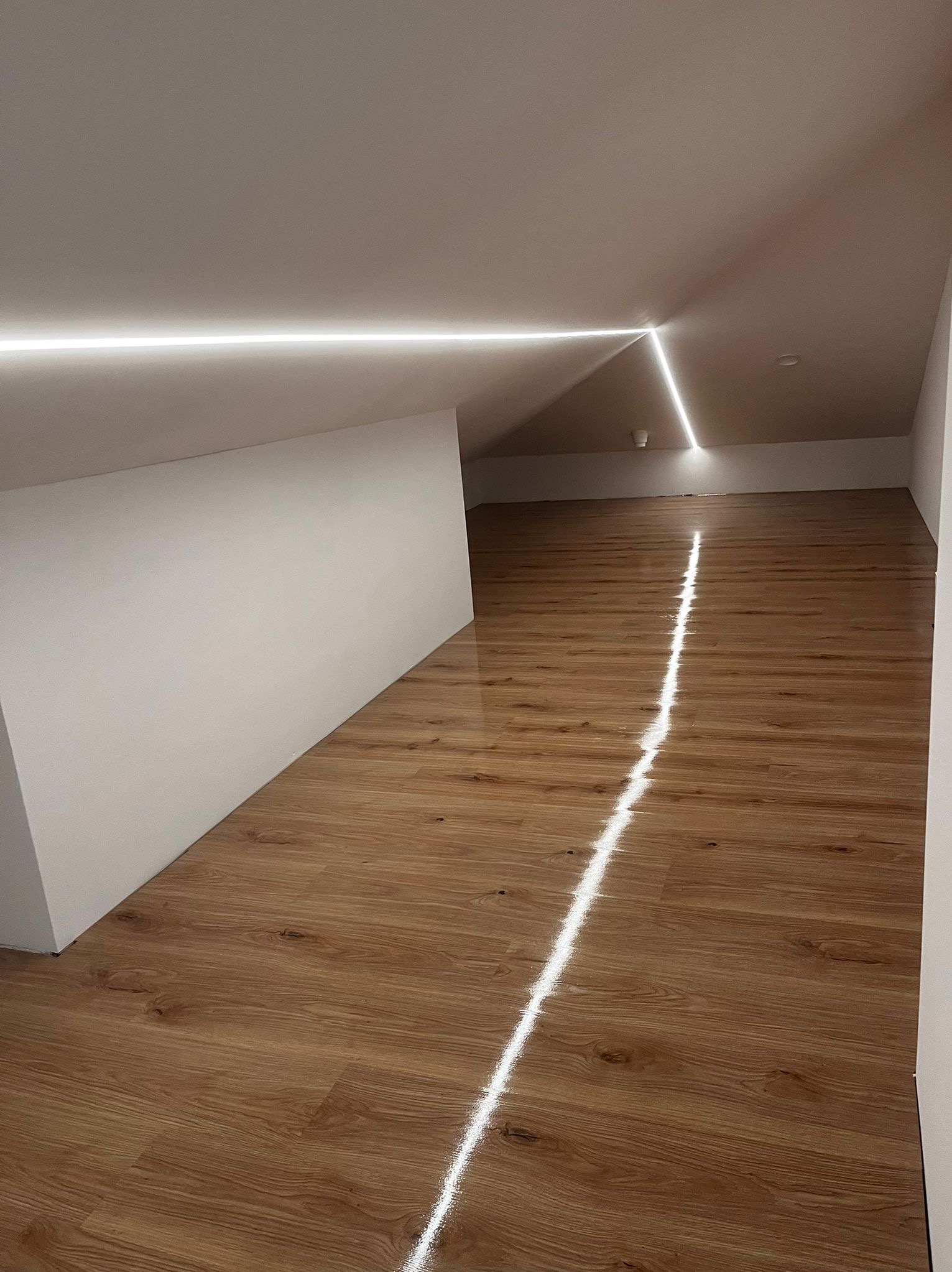 Jasne panele podłogowe w nowym poddaszu ze skośnym sufitem i nowoczesnym oświetleniem LED wzdłuż krawędzi sufitu. Minimalistyczny design.