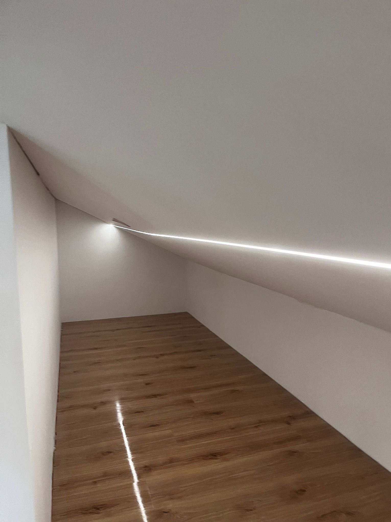 Wykończone poddasze ze skośnym sufitem, drewnianą podłogą i subtelnym oświetleniem LED wzdłuż krawędzi sufitu. Minimalistyczny design, jasne ściany.