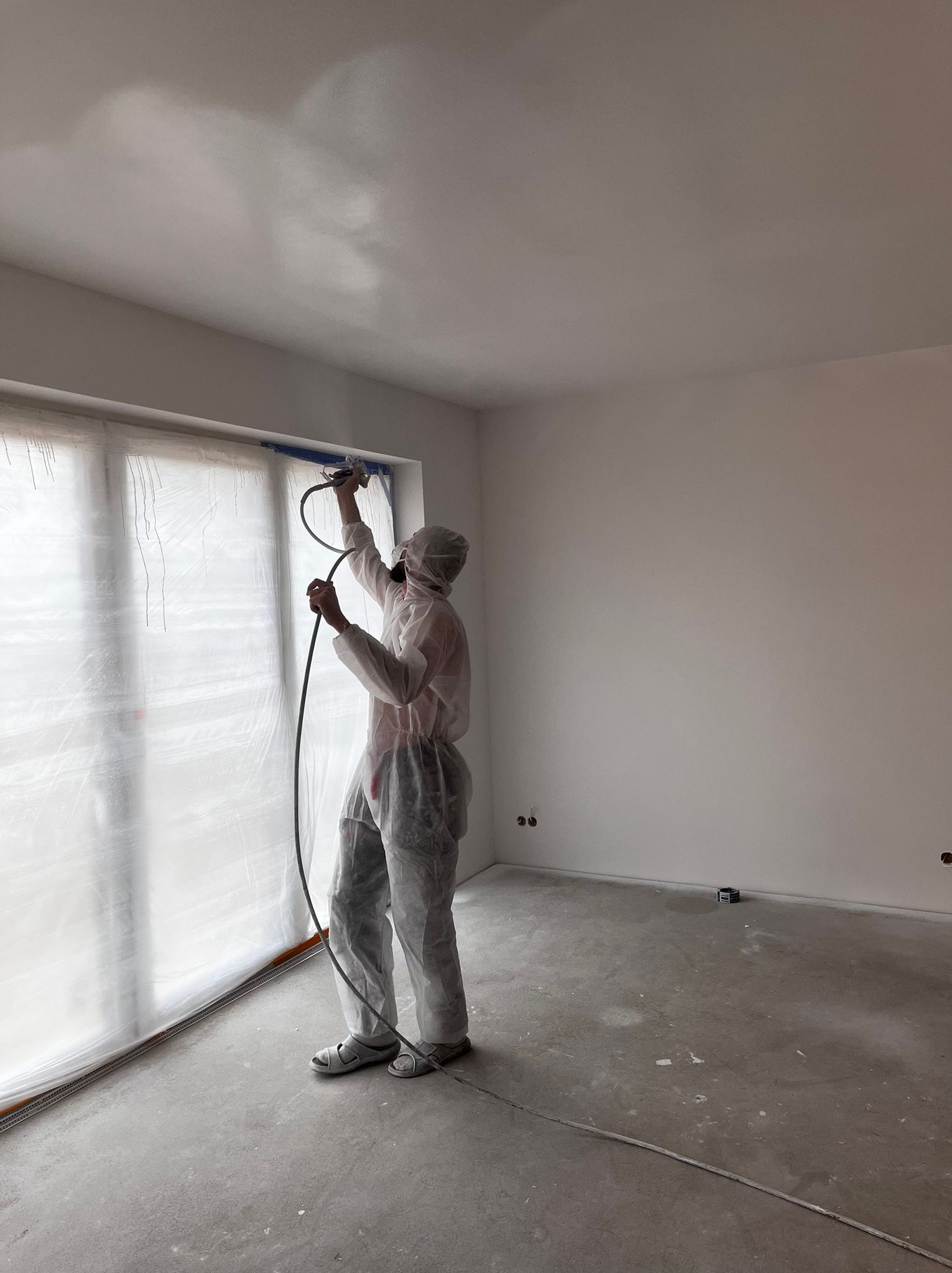 Pracownik w kombinezonie ochronnym maluje ściany i sufit w pomieszczeniu za pomocą pistoletu natryskowego. Okno zabezpieczone folią malarską.