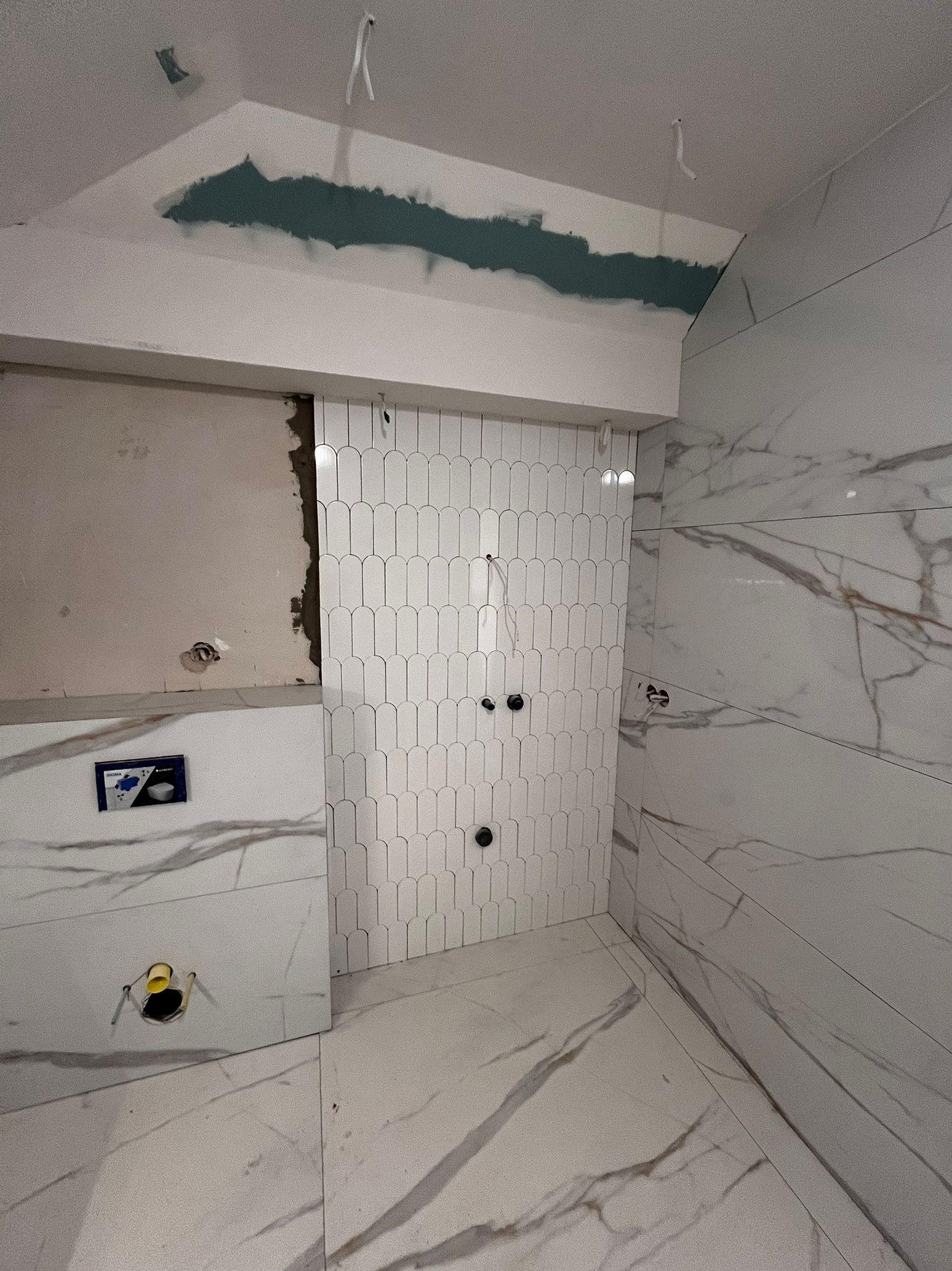 Białe, podłużne płytki ułożone w pionowy wzór na ścianie łazienki, kontrastujące z marmurowymi płytami. Widoczne przygotowania do montażu armatury.