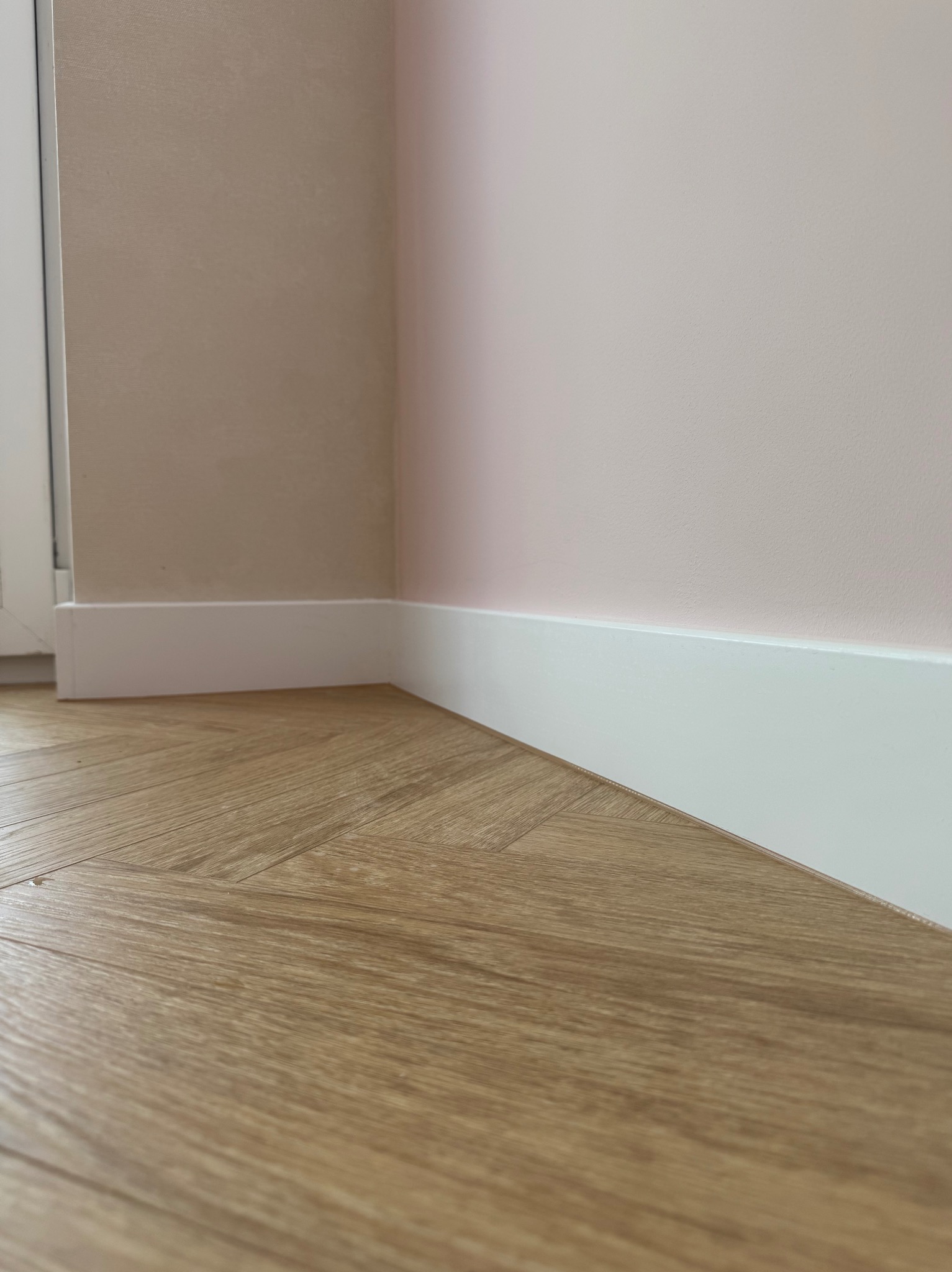 Jasne panele ułożone w jodełkę, białe listwy przypodłogowe przy ścianach w odcieniach beżu i różu. Minimalistyczne wykończenie wnętrza, detale podłogi i ścian.