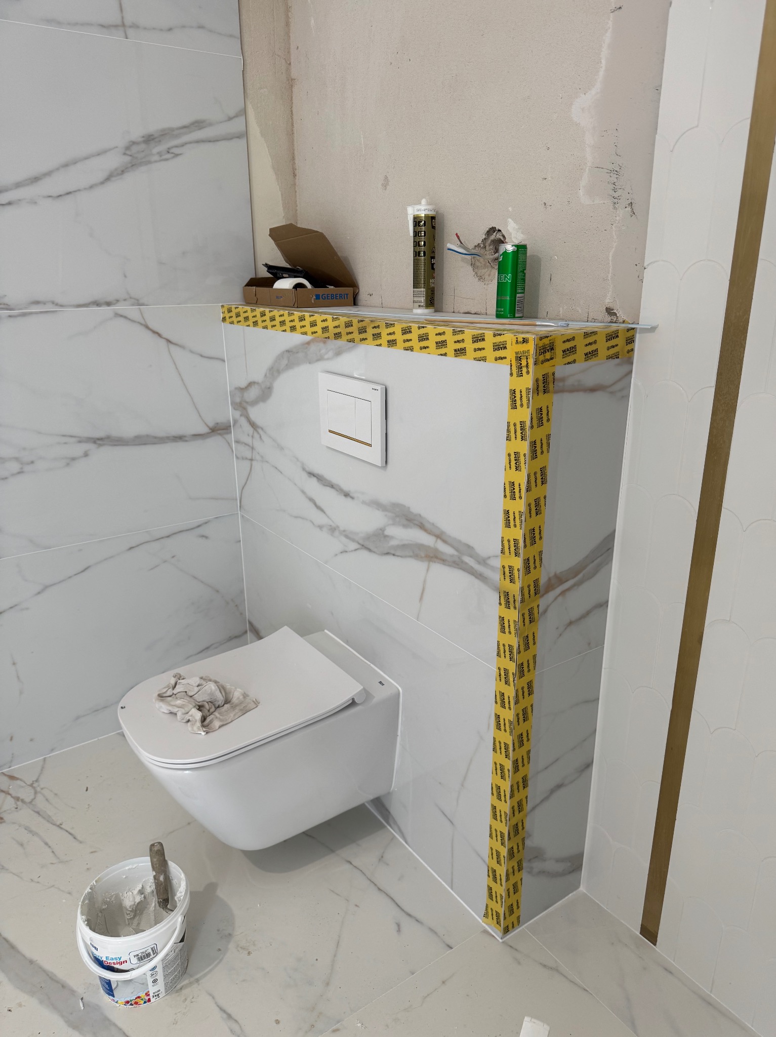 Biała, wisząca toaleta w trakcie montażu w łazience z marmurowymi płytkami. Widoczne narzędzia i materiały budowlane, taśma zabezpieczająca. Ujęcie z dołu.