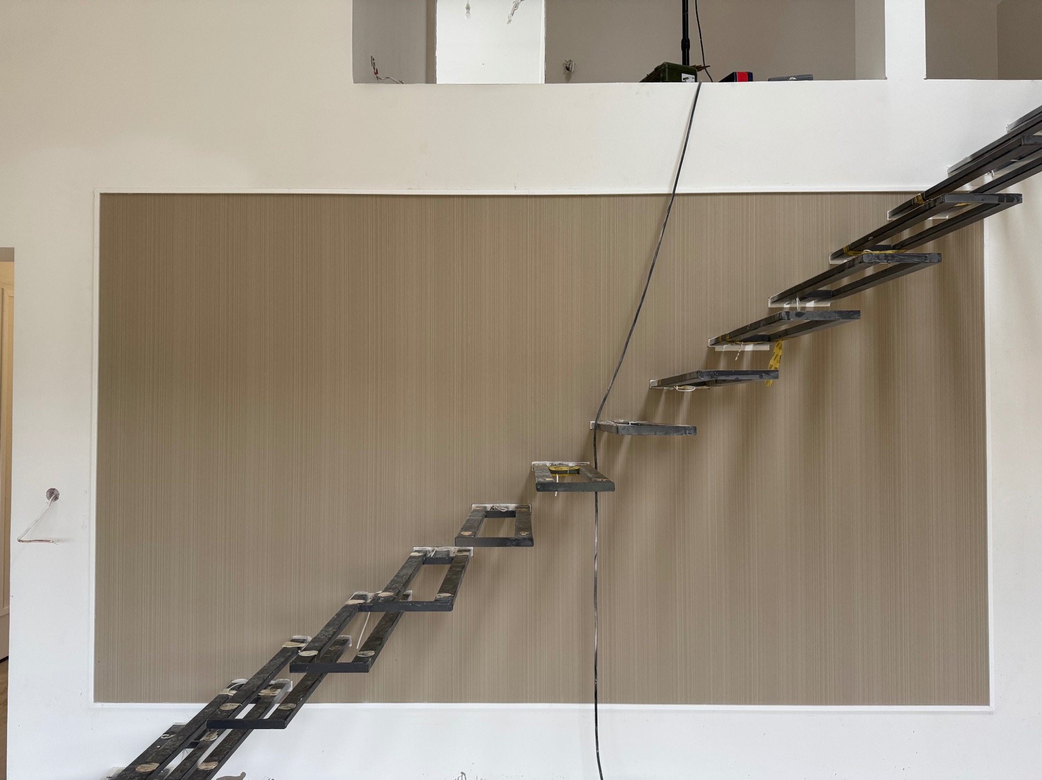 Montaż ażurowych schodów metalowych na ścianie z tapetą w odcieniach beżu. Widoczne elementy mocujące i przewody. Etap prac wykończeniowych w jasnym wnętrzu.