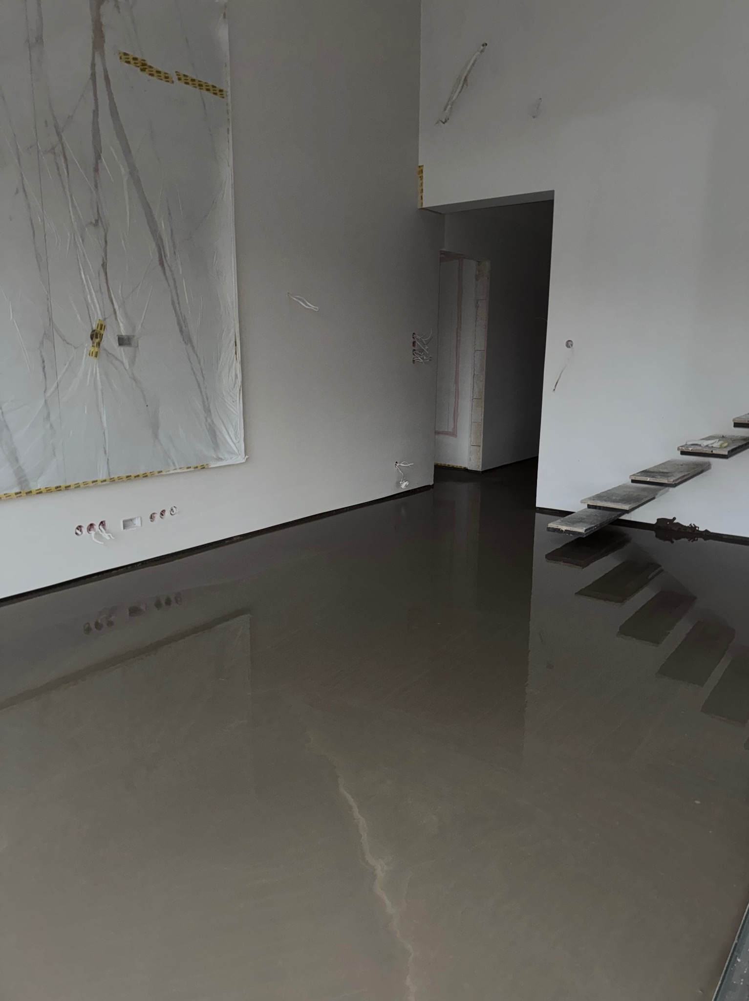 Wnętrze w trakcie prac: wylany, mokry podkład podłogowy, zabezpieczona folią ściana, widoczne schody. Surowe wykończenie, przygotowanie do dalszych prac.