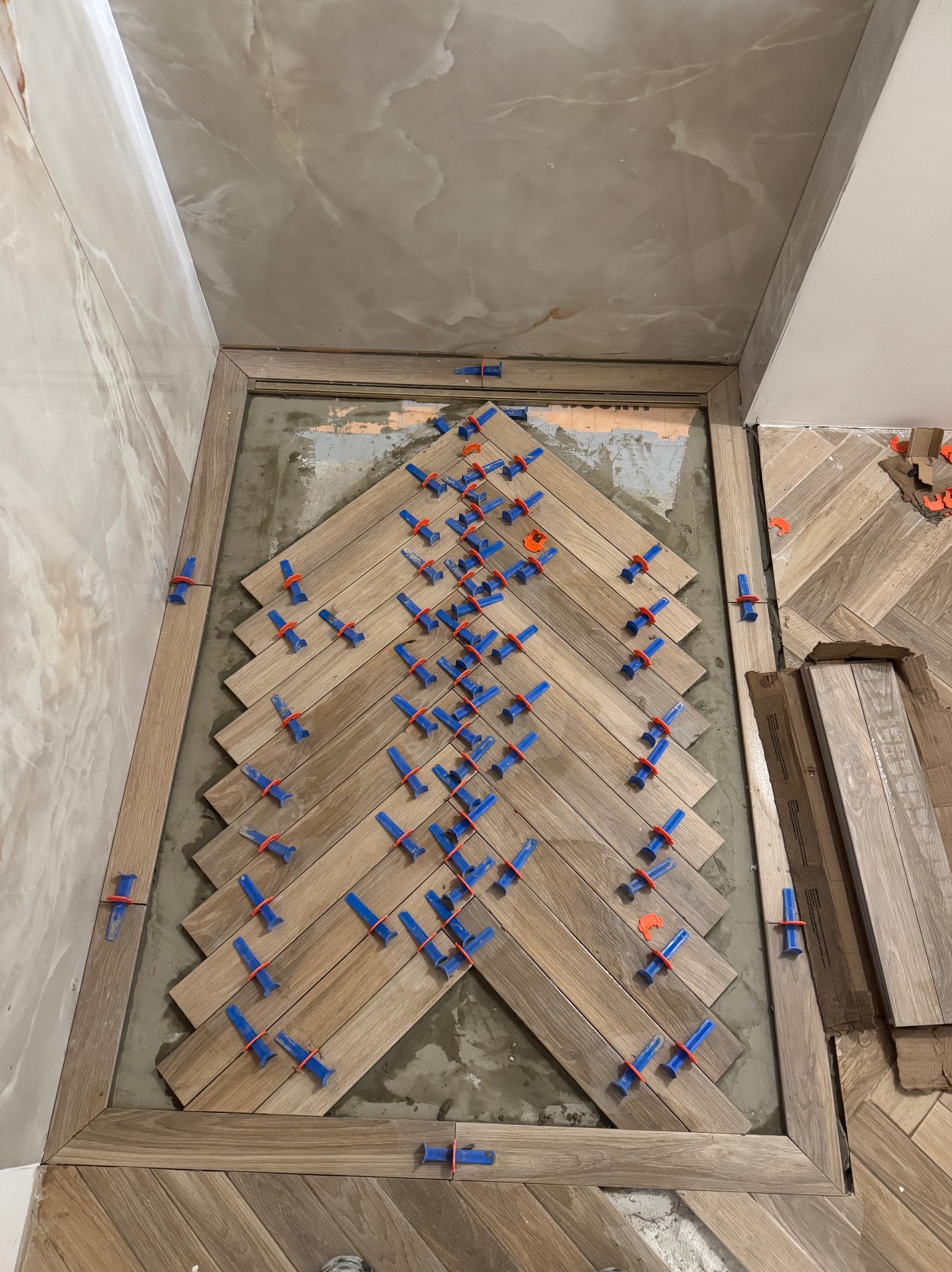 Układanie parkietu w jodełkę z użyciem klipsów poziomujących. Widoczne docięte deski i fragment wykończonej podłogi. Jasne drewno, ciepłe oświetlenie.