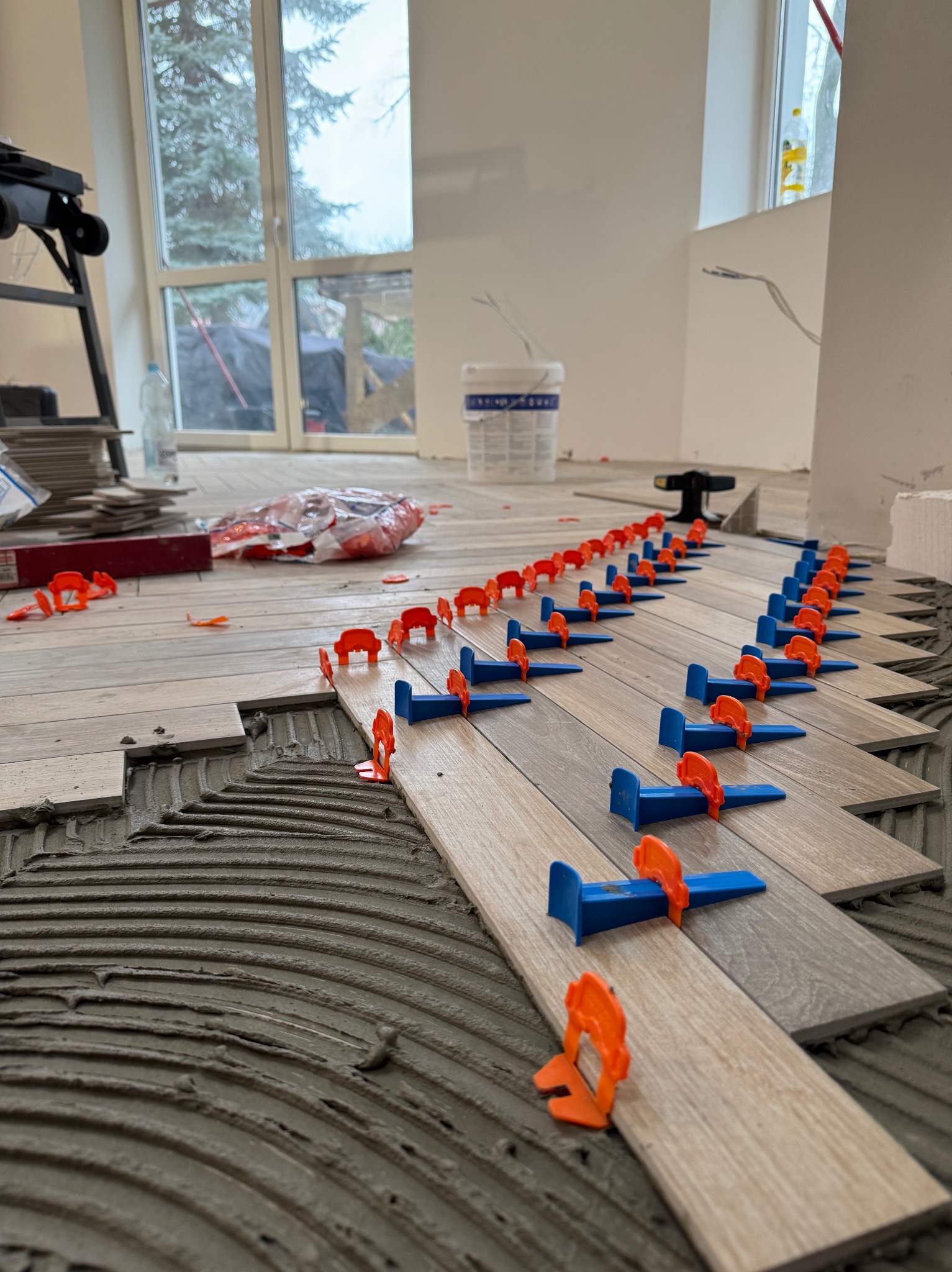 Układanie płytek podłogowych z użyciem systemu poziomowania. Widoczne klej, płytki imitujące drewno i klipsy. W tle okno i materiały budowlane.