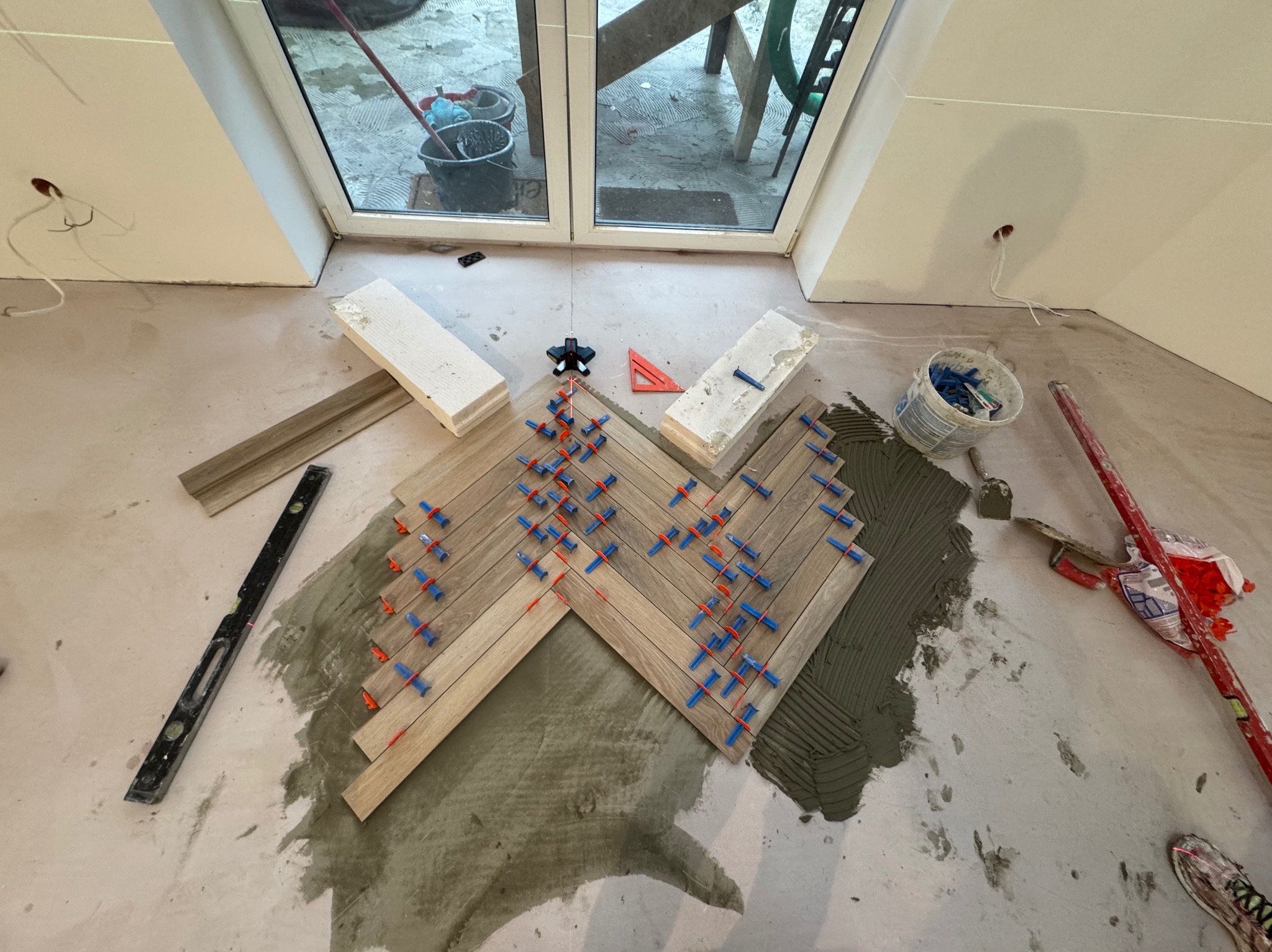 Układanie parkietu w jodełkę na podłodze pod skosem, z użyciem poziomicy laserowej i klipsów dystansowych. Widoczne narzędzia i materiały budowlane.