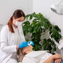 ATELIER UŚMIECHU SPÓŁKA Z OGRANICZONĄ ODPOWIEDZIALNOŚCIĄ - Stomatolog w białym fartuchu i niebieskich rękawiczkach podczas badania jamy ustnej pacjentki leżącej na fotelu dentystycznym; w tle roślina doniczkowa i lampa dentystyczna.
