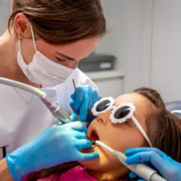 Lekarz w masce i niebieskich rękawiczkach wykonuje zabieg stomatologiczny u dziecka w fotelu dentystycznym, dziecko ma na oczach okulary w kształcie kwiatków.