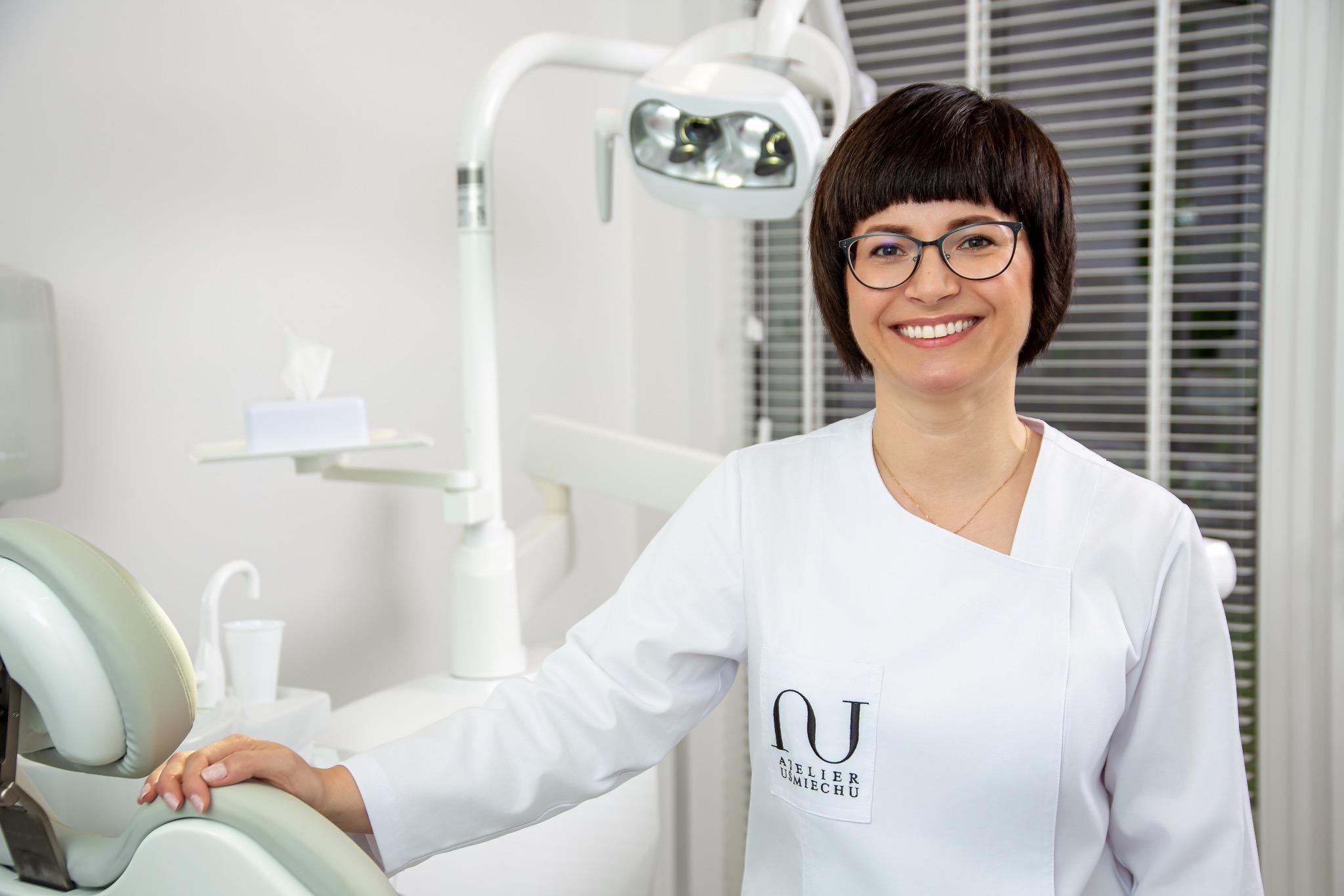Uśmiechnięta kobieta w okularach i białym fartuchu lekarskim, stojąca w gabinecie stomatologicznym z widocznym fotelem i lampą.