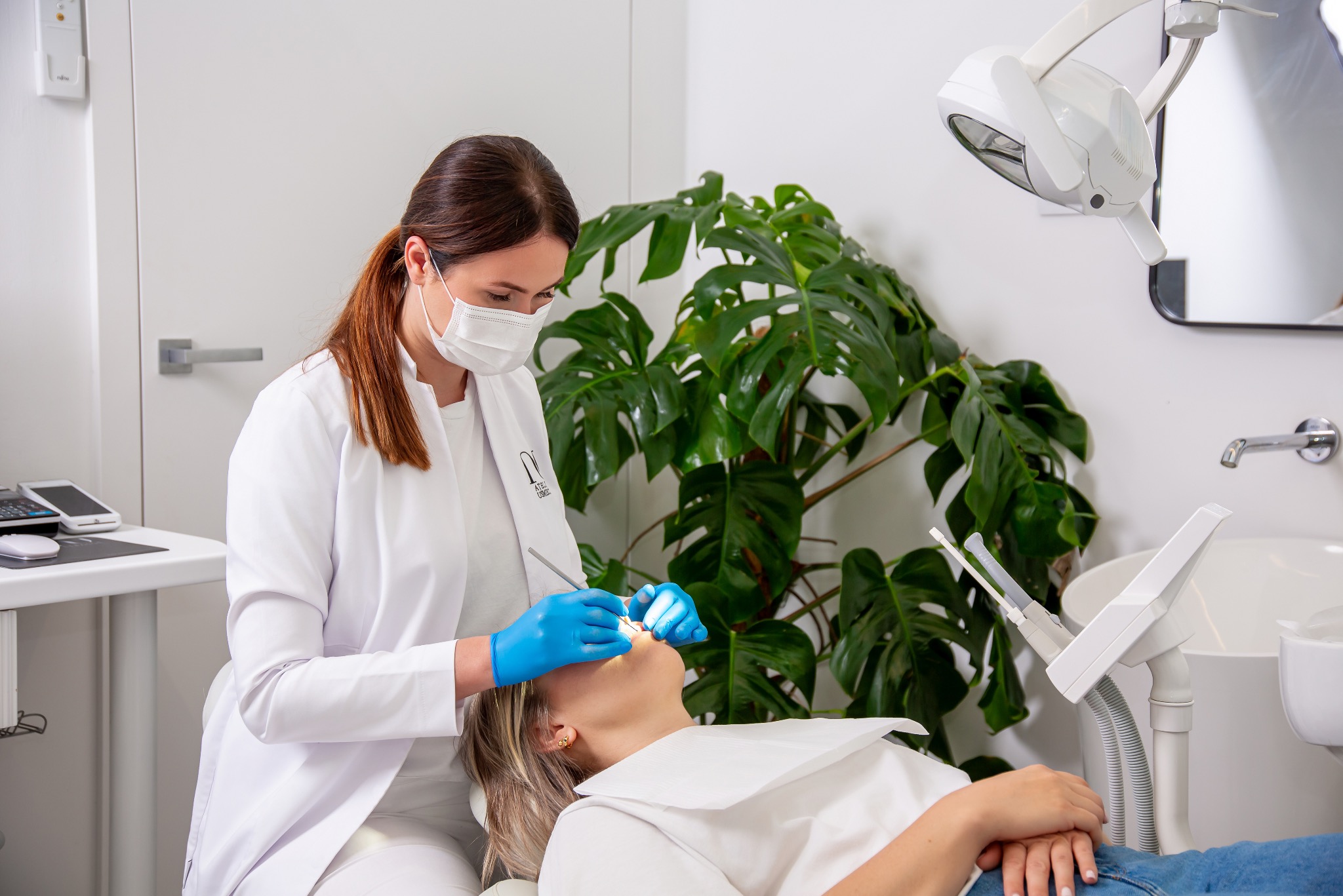 Stomatolog w białym fartuchu i niebieskich rękawiczkach podczas badania jamy ustnej pacjentki leżącej na fotelu dentystycznym; w tle roślina doniczkowa i lampa dentystyczna.