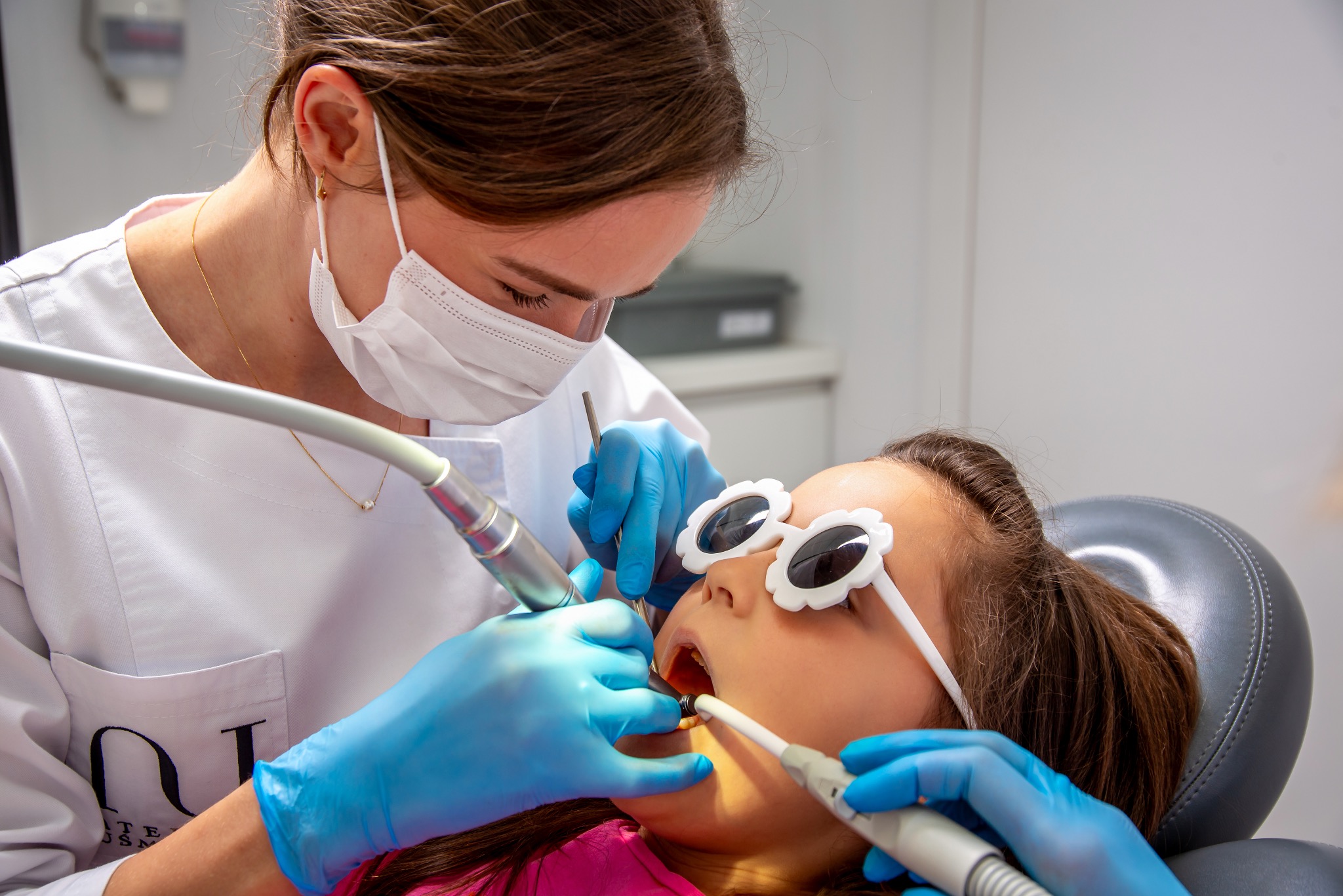 Lekarz w masce i niebieskich rękawiczkach wykonuje zabieg stomatologiczny u dziecka w fotelu dentystycznym, dziecko ma na oczach okulary w kształcie kwiatków.