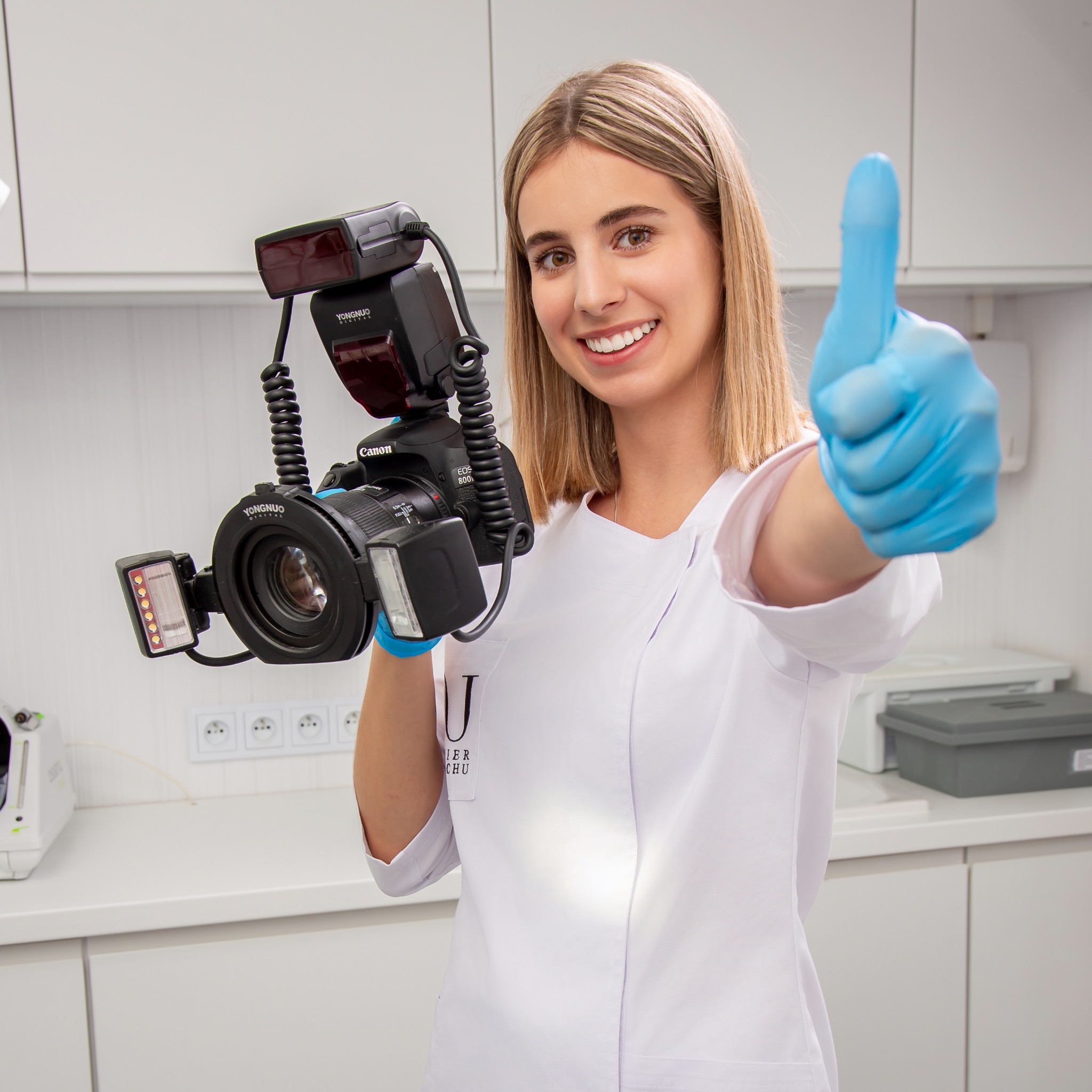 Uśmiechnięta kobieta w białym uniformie i niebieskich rękawiczkach trzyma aparat Canon EOS 80D z lampami błyskowymi Yongnuo, pokazując kciuk w górę w gabinecie z białymi szafkami.